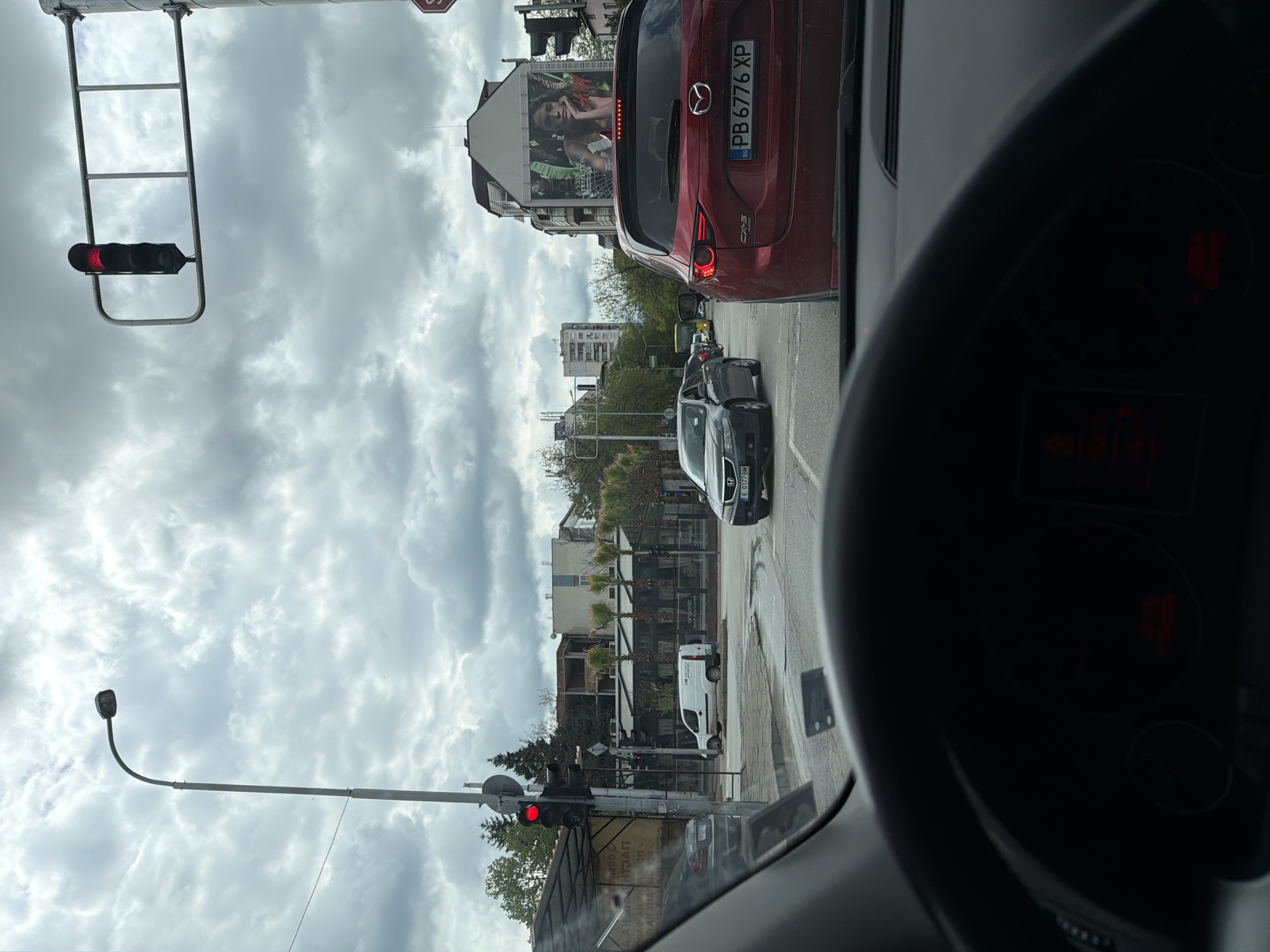 The image captures a street view from inside a vehicle, stopped in traffic on an overcast day in Plovdiv, Bulgaria. A red traffic light is visible on the left, indicating the halt.
In the foreground, parts of the car's steering wheel and dashboard are visible, with faint red illuminated displays. Directly ahead, a red Mazda CX-5 SUV is stopped, displaying the Bulgarian license plate "PB 6776 XP," which is consistent with Plovdiv registration. Further down the street, a black sedan with the license plate "K 0177 BM" (Sofia registration) and a white delivery van are also part of the traffic.
The sky is heavily clouded, suggesting an overcast daytime. Along the street, several multi-story buildings are visible. One prominent building features a large billboard with an image of a woman and partially readable English text such as "A STORY" and "NATURAL." Other commercial establishments along the street display Cyrillic text, including "ЧАФАН ЯНАЯ," "AAHNTPAΠ," "BEL MOVHЕ НУ," and "organic." Several palm trees and other greenery are scattered among the buildings.