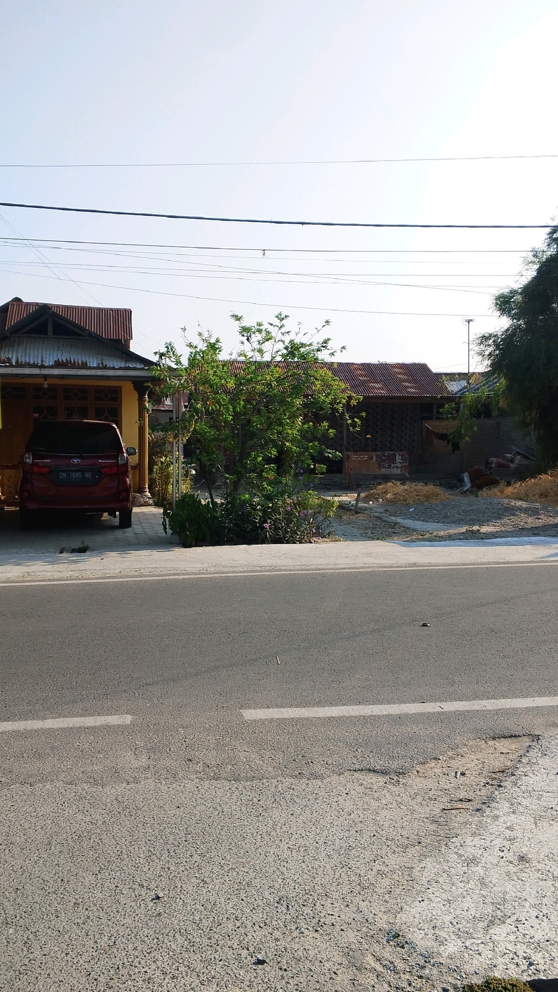 A red minivan is parked in the driveway of a yellow house with a corrugated metal roof. The house is located on a street in Talise, Indonesia, with a paved road in the foreground and power lines overhead. A leafy green tree stands in front of the house, and a second, darker building with a similar roof is visible behind it. The area appears to be in a rural or suburban setting.