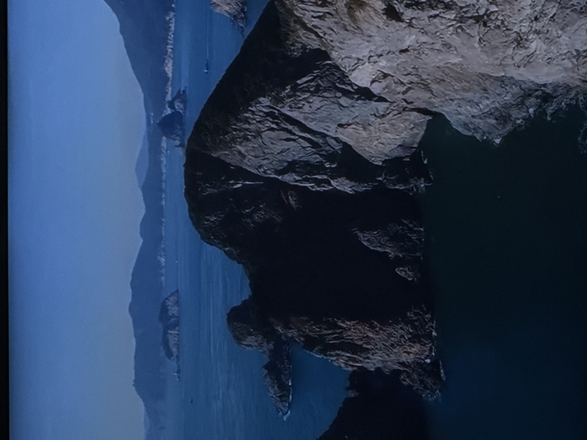 The image displays a serene, coastal landscape dominated by rugged, dark brown rock formations jutting out of the deep blue sea. The formations are jagged and textured, with visible cracks and crevices, suggesting a weathered and ancient appearance. In the background, several smaller islands and mountain ranges are faintly visible, shrouded in a soft, atmospheric haze. The sky is a clear, deep blue, transitioning into a lighter shade near the horizon. The overall scene evokes a sense of natural beauty and isolation, with no visible signs of human presence or activity. The location is identified as the city of Sinyavino, Russia.
