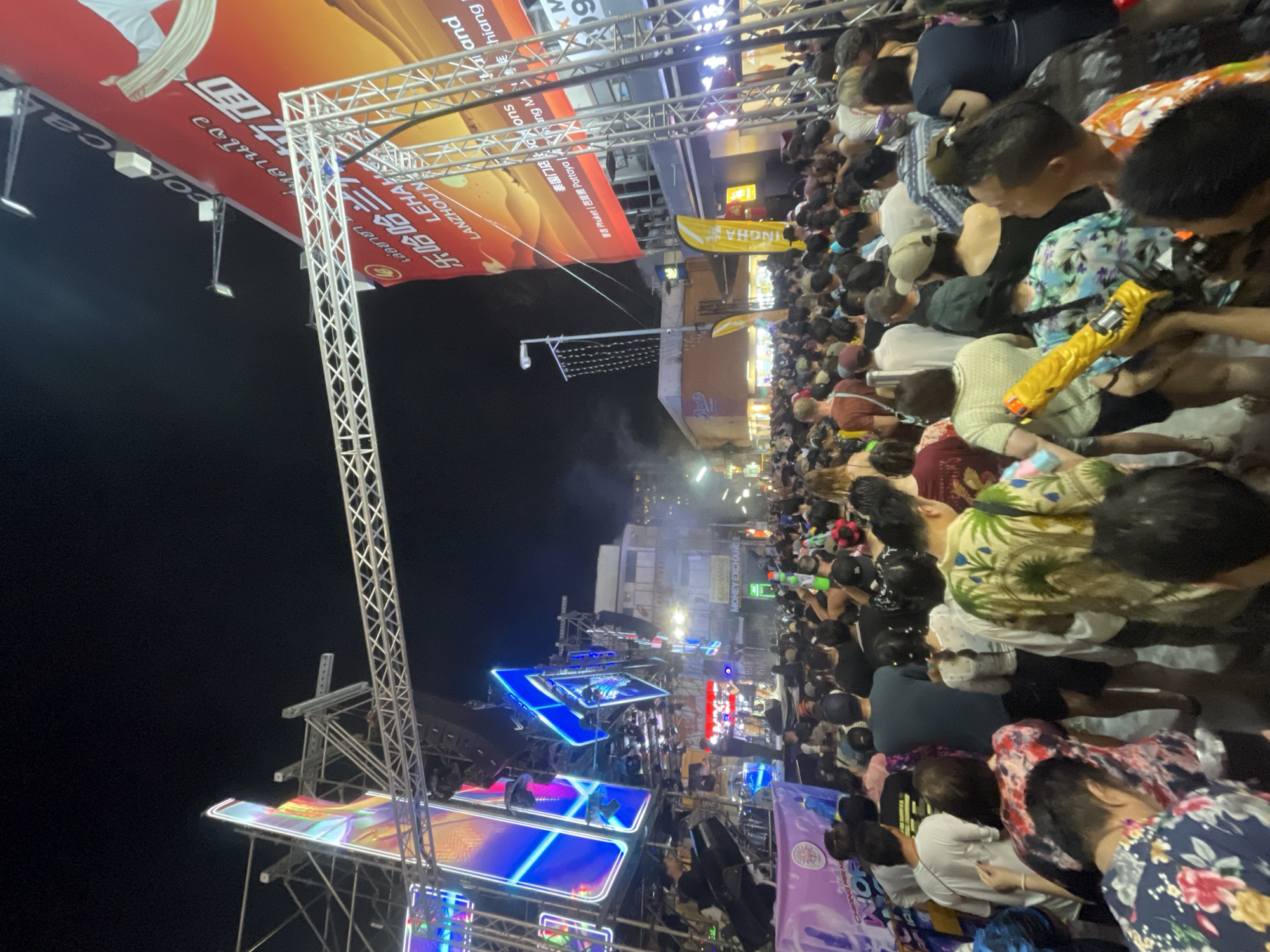 This image depicts a large crowd of people gathered at night in what appears to be a celebration or event. The location seems to be in the Chiang Mai City Municipality, Thailand, as indicated by signs displaying place names like "Chiang Mai" and Thai script. Many people are holding water guns, suggesting a water festival or celebration. The presence of a large stage structure with lighting indicates a performance or event space. Signs with the brand "LEHA LEHAO" and phrases in Chinese are visible. People are dressed in various casual clothing, including floral shirts. The overall environment is lively and crowded, typical of a festival or public event.
