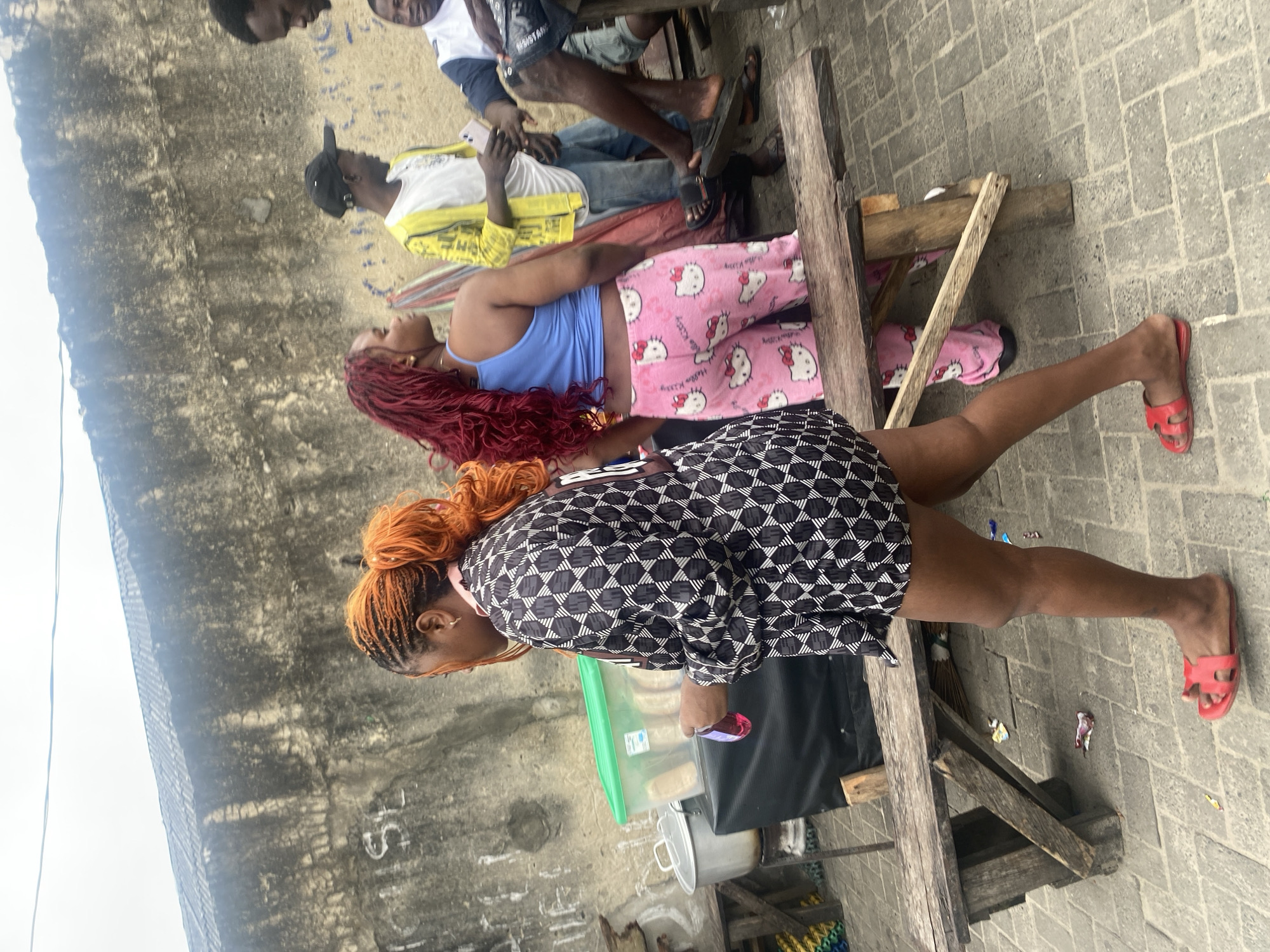 In a gritty urban setting, an outdoor market scene unfolds under an overcast sky. A woman with vibrant orange braided hair, wearing a patterned black and white dress, walks across a cobblestone path. She carries a pink bottle in her left hand. To her right, another woman with long, wavy red hair, wearing a blue tank top and pink Hello Kitty patterned pants, stands looking away from the camera. Behind them, a man in a yellow jacket and white t-shirt is visible, holding a smartphone. Another man in a blue and white shirt and shorts sits on a wooden bench, partially obscured. The background features a weathered concrete wall with graffiti and a stall with cooking equipment. The overall impression is one of daily life in a bustling, possibly developing, urban environment.