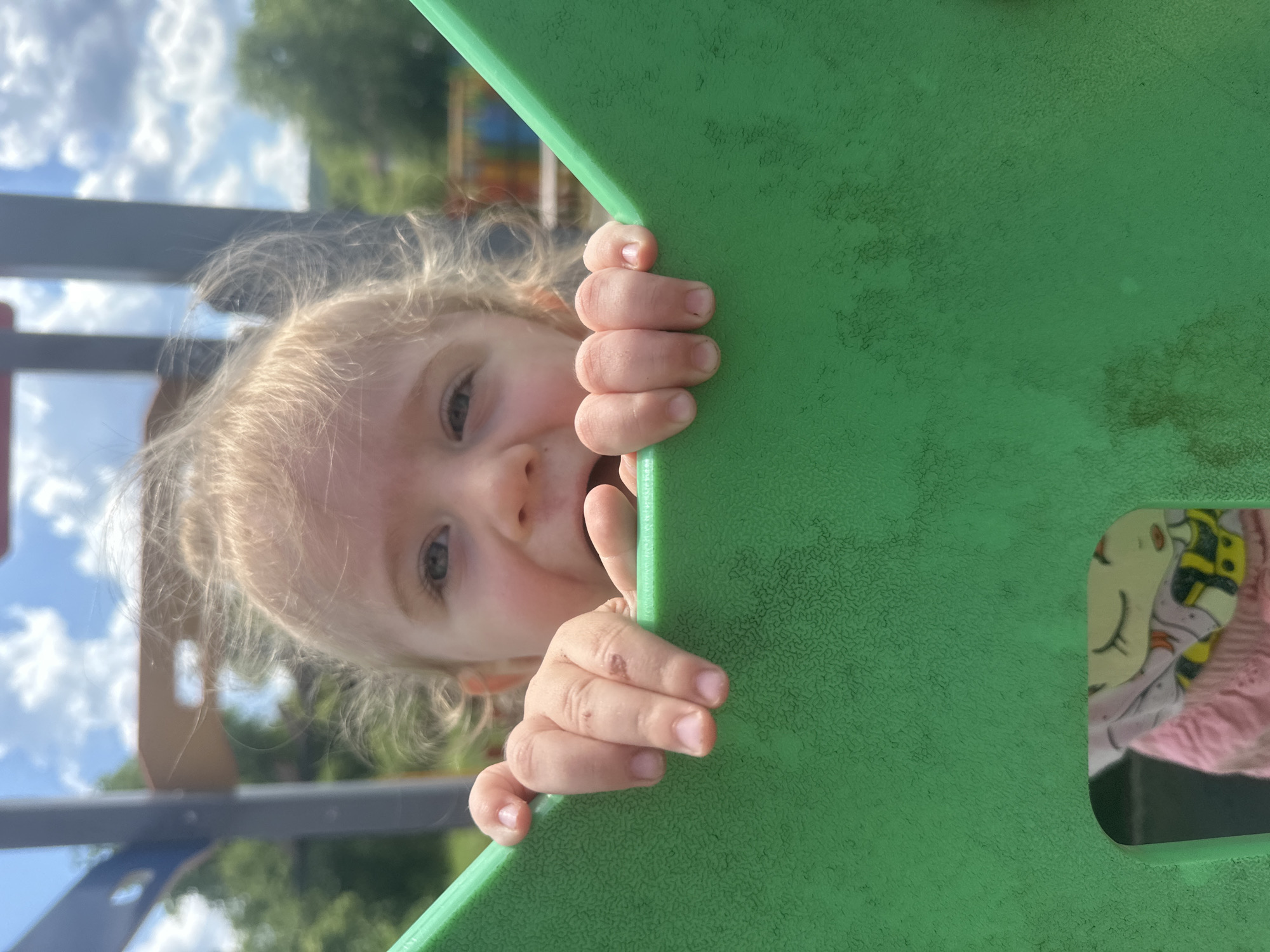 In Donchovci, Bulgaria, a close-up shot captures a young child peeking over a bright green plastic structure, possibly part of a playground. The child, with light-colored, wispy hair, looks directly at the camera with a wide-eyed expression. Their small hands, with visible fingernails and minor abrasions on one, grip the edge of the green structure. The background is blurred, showing a bright sky with scattered clouds and hints of green trees and playground equipment, suggesting an outdoor setting on a sunny day. A glimpse of another child's clothing, featuring a cartoon character and pink fabric, is visible through an opening in the green structure.