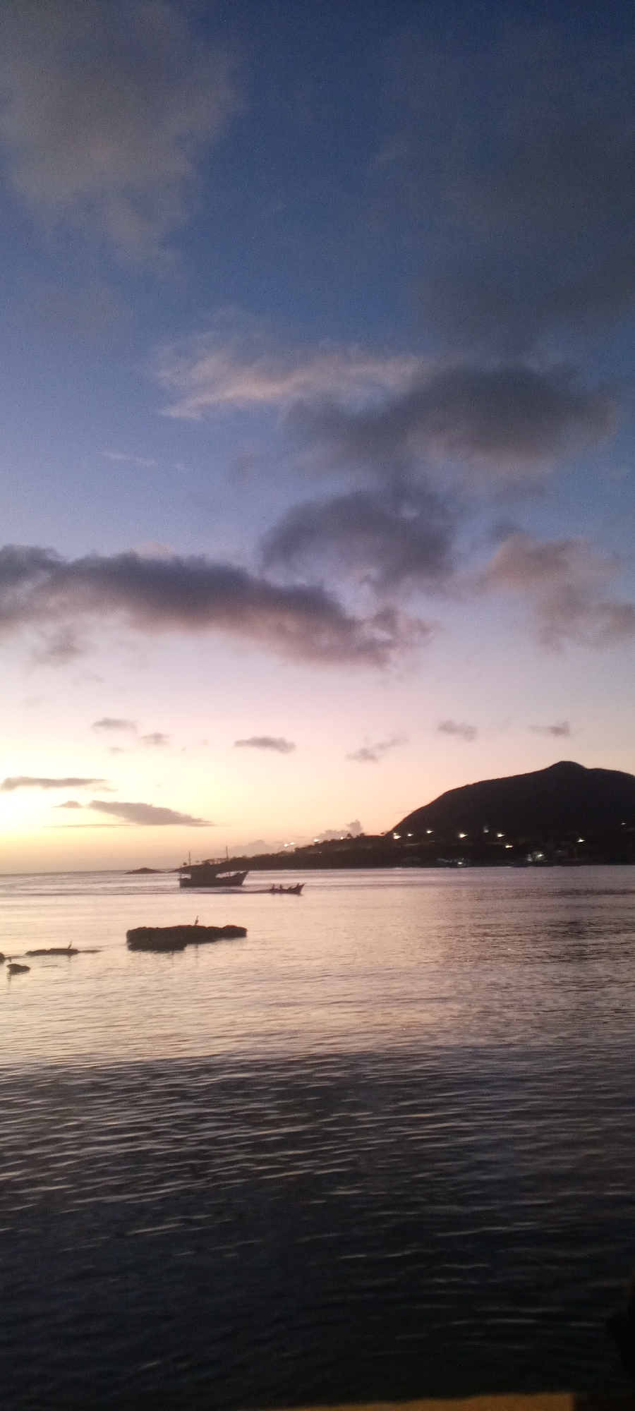 The image shows a serene coastal scene at dusk in Juan Griego, Venezuela. The sky is a gradient of deep blue at the top, transitioning to soft purples and pinks near the horizon, with scattered clouds tinged by the setting sun. The calm water of the bay reflects the colors of the sky, creating a muted, shimmering surface. In the middle ground, several boats are visible on the water. A larger, silhouette of a boat with a distinctive sail is moving from left to right. Behind it, a smaller boat with multiple occupants is also in motion. In the foreground, dark, rocky formations are partially submerged in the water, with a bird perched on one of them. The background features a dark, silhouetted mountain range that overlooks the bay. Lights from what appears to be a town or settlement are scattered along the coastline at the base of the mountains, indicating human presence. The overall atmosphere is peaceful and tranquil.