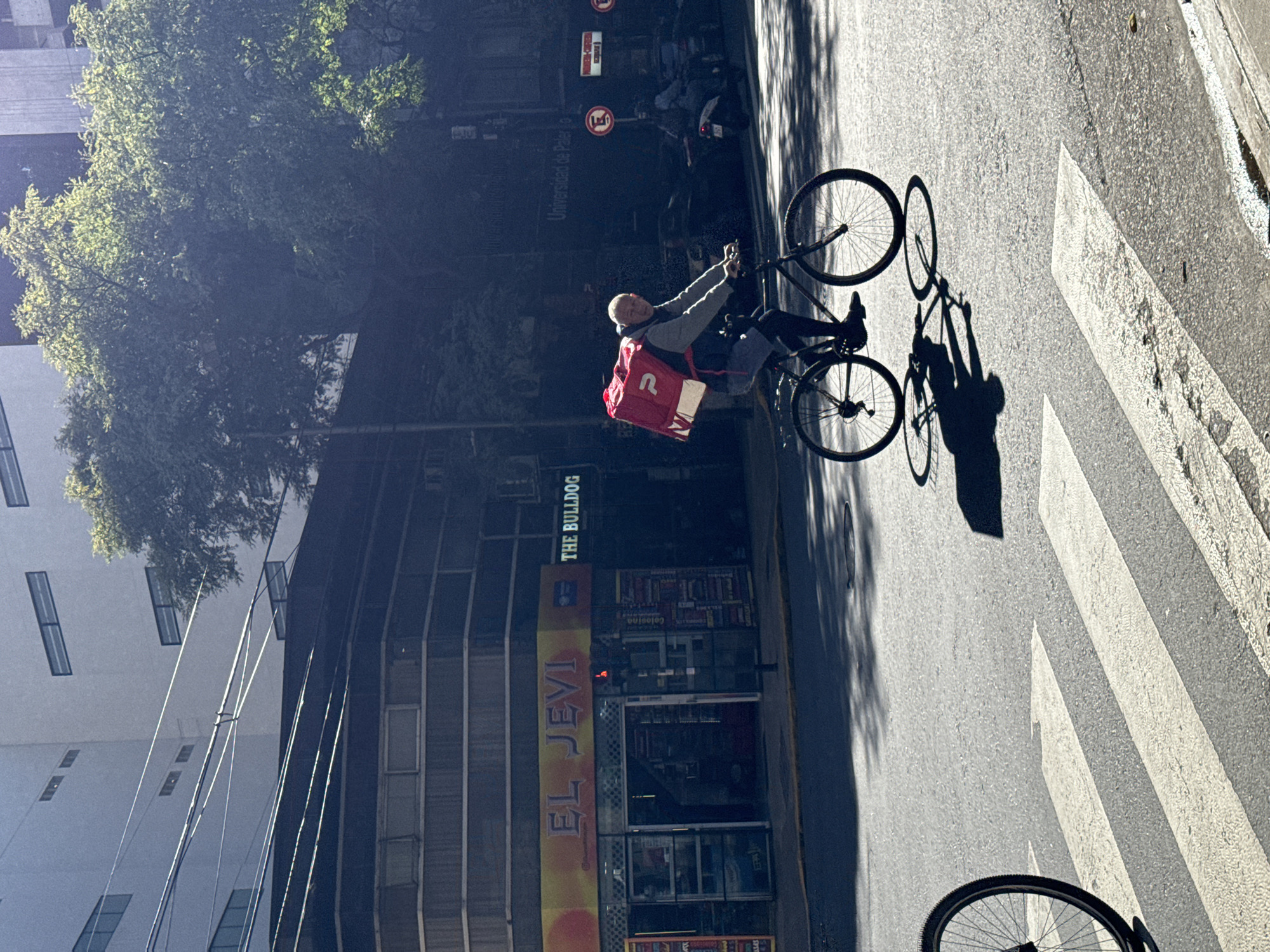 A delivery cyclist is riding a bicycle across a crosswalk. He wears a red delivery backpack and grey jacket. The cyclist is in front of a building with two storefronts. One store is called "EL JEVI" and the other store is "THE BULLDOG". A sign that reads "Universidad de Palerm" is visible. The bottom of the photograph shows the wheel of another bicycle. The location is Buenos Aires, Argentina.