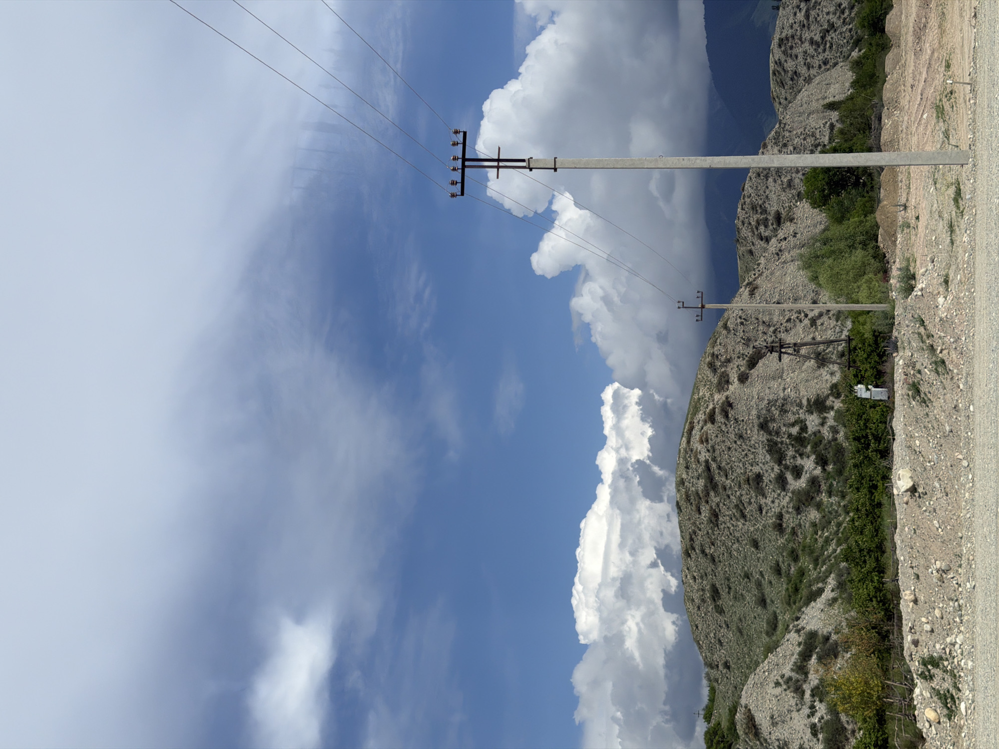 The image depicts a scenic view of Tamga, Kyrgyzstan, featuring rolling hills covered in sparse vegetation and gravel. A clear blue sky with scattered white clouds dominates the upper half of the frame. In the foreground, a concrete power pole with wires stretches diagonally across the image. Another, smaller power pole is visible in the mid-ground. The hills are characterized by their rocky terrain and patches of green shrubs. The overall scene conveys a sense of rural tranquility and natural beauty.