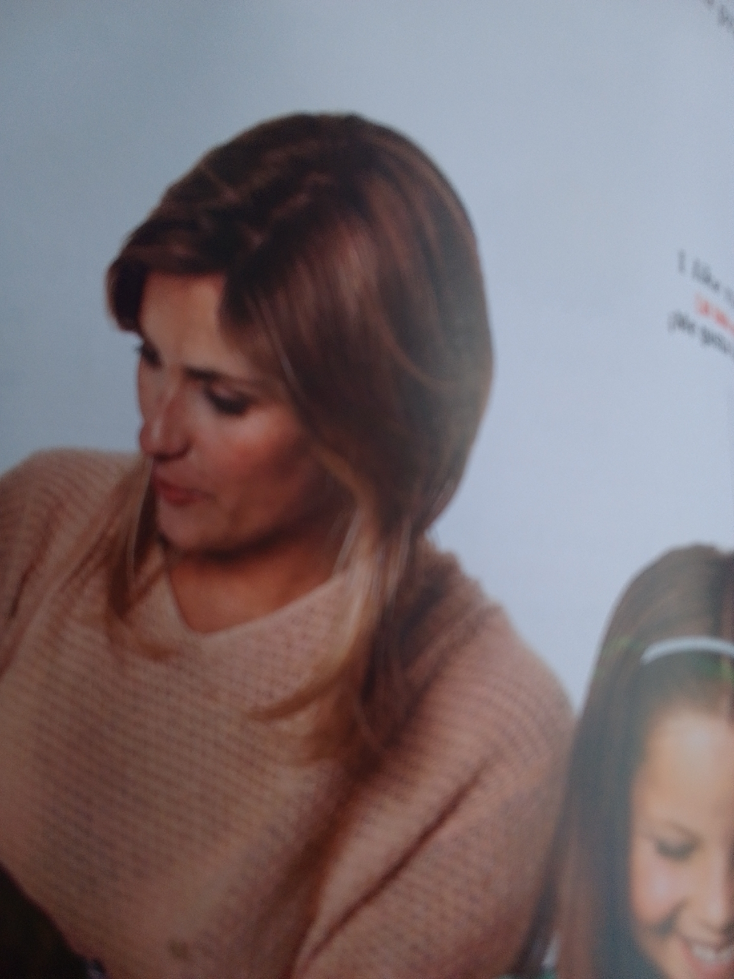 The media file shows a medium shot of two people, an adult woman and a young girl, in what appears to be an indoor setting. The woman, positioned on the left, has light skin, brown hair styled in a braid, and is wearing a tan sweater. Her gaze is directed downwards and to the side, with a neutral expression. To the right, the young girl, also with light skin, is partially visible. She has brown hair and is wearing a white headband. Her face is turned towards the right, and she appears to be smiling. The background is a plain, light gray, suggesting an uncluttered environment. There is some faint, blurry text visible on the right side of the image, but it is illegible. No specific time of day, weather conditions, or strong location cues are discernible. The overall mood seems to be calm and perhaps intimate, given the closeness of the subjects.
