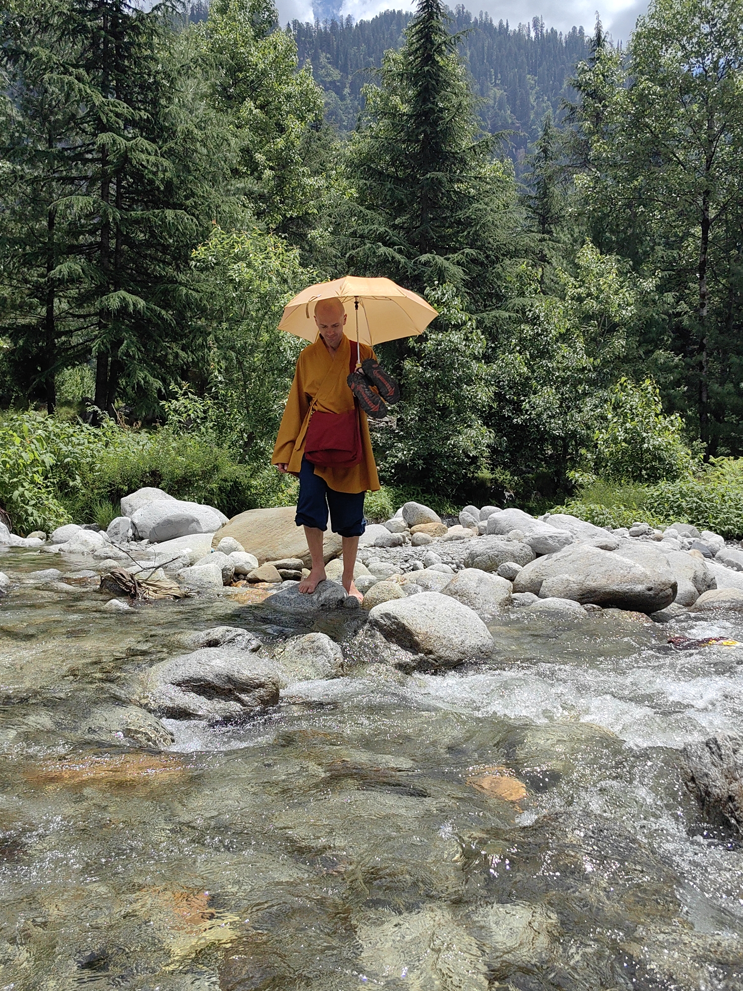 A bald, light-skinned man wearing a yellow robe and dark blue shorts stands in a shallow, rocky river in Kulang, India. He is holding a light orange umbrella above his head and has a red bag slung over his right shoulder. The man is barefoot and appears to be walking on stones in the river. The riverbed is composed of numerous gray and tan rocks, some of which are submerged in the clear, flowing water. Lush green trees and foliage surround the riverbank, with more trees extending up a steep, forested mountainside in the background. The sky is partly cloudy with patches of blue visible.