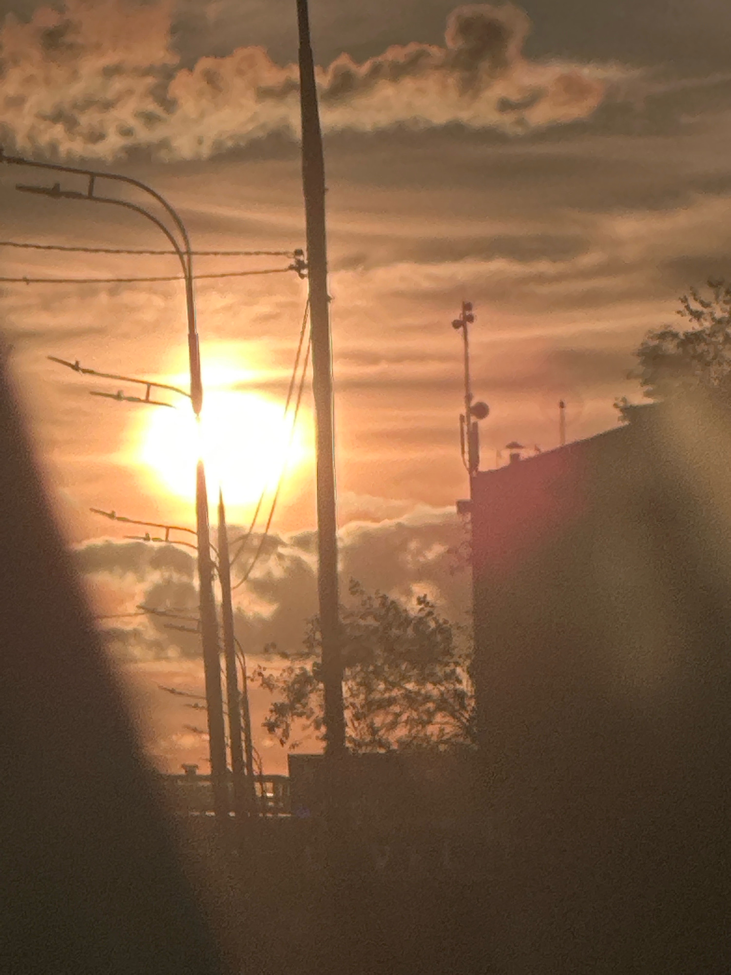 This image depicts a sunset over a city, with a bright sun partially obscured by clouds. Power lines and utility poles are visible in the foreground, silhouetted against the sky. The sun's rays cast a warm, orange glow across the scene, illuminating the clouds and the tops of buildings in the background. The overall impression is one of a serene, urban landscape at dusk.