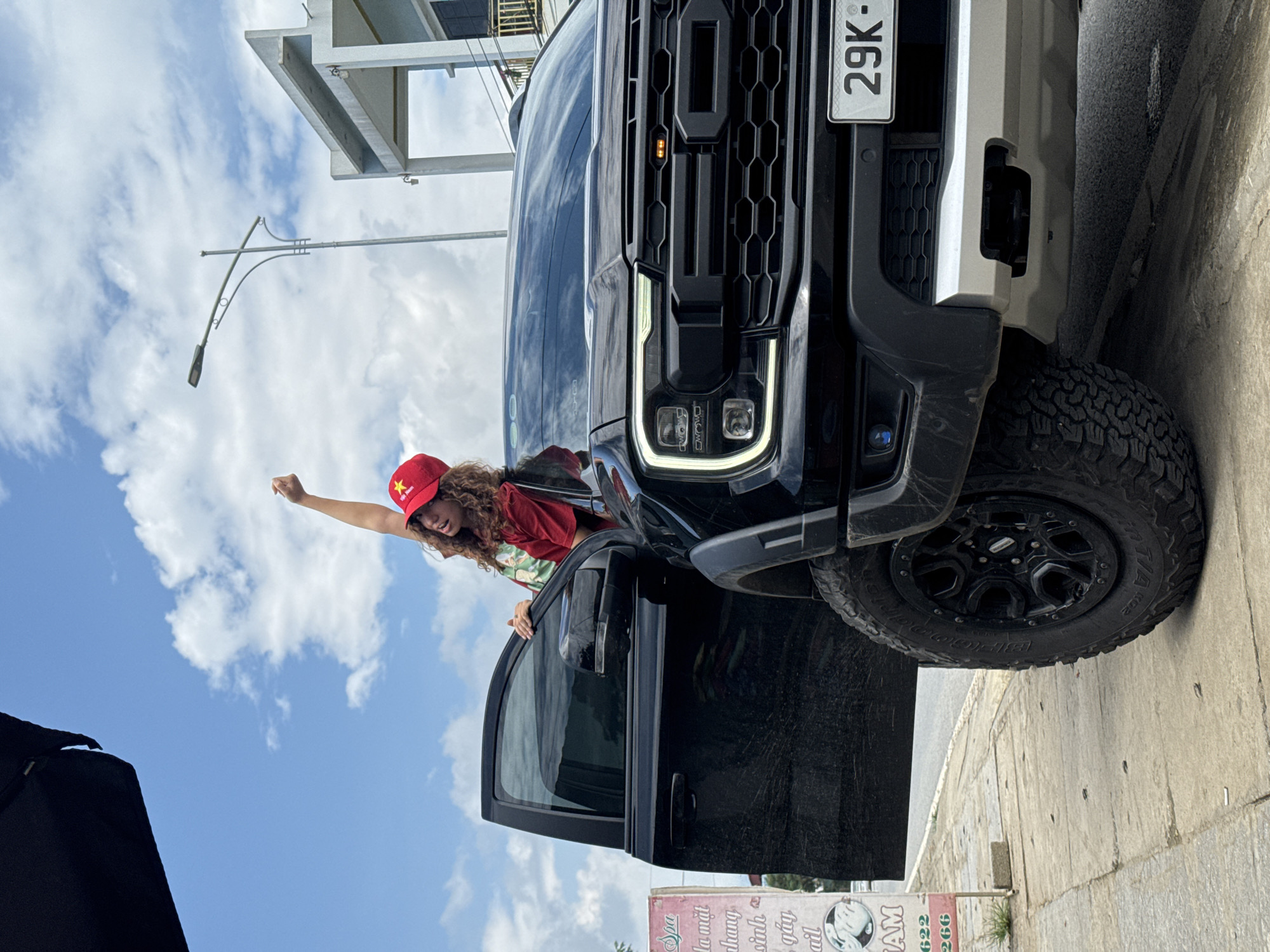 The image shows a woman in a red cap and shirt standing next to the open door of a black Ford Raptor truck. The truck's license plate reads "29K-". The woman has curly hair and her arm is raised in a fist. The background includes a building, a street light, and a cloudy blue sky. A sign at the bottom of the image reads "Spa la mat hang sinh gay ail AM 622 266". The location appears to be in the city of Trung Tam Ward, Vietnam.