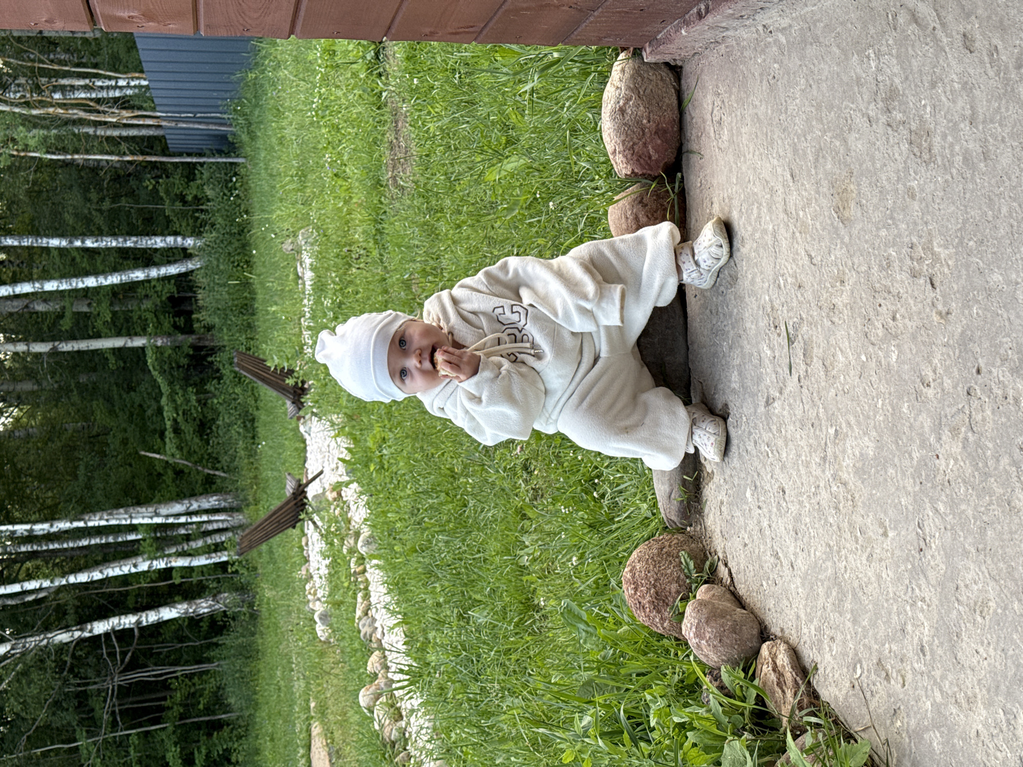 A young child, wearing a white hooded jumpsuit and matching hat, sits on a rock outdoors, with a mouthful of food. The child has blue eyes and is looking directly at the camera. The background consists of a grassy area with trees, a wooden fence, and part of a wooden building. The child appears to be in a rural or semi-rural setting. The ground in front of the child is paved with concrete. The image was taken in Ryzhkovo, Russia.