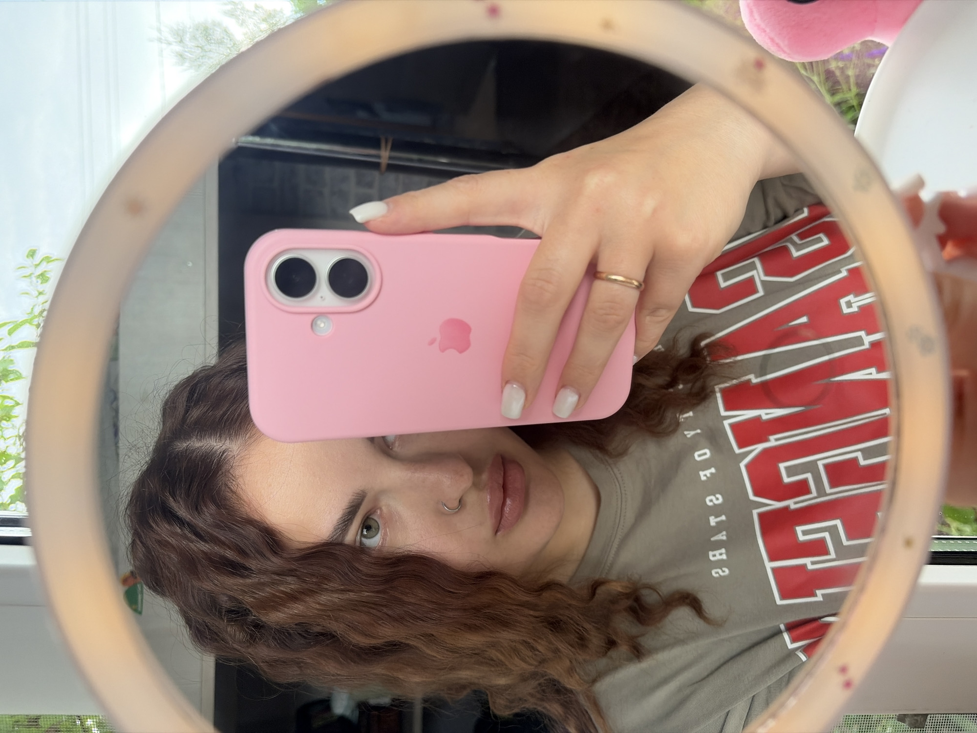 A person with brown curly hair is taking a selfie using a pink mobile phone with two camera lenses and an Apple logo on the back. The person's left hand is holding the phone and has a gold ring on the ring finger. The person has light skin, a nose ring, and painted nails. The reflection is captured in a ring light. The person is wearing a gray t-shirt with large red letters that are partially mirrored, spelling out "JERSEY." Outside the window, there are green plants. A pink plush toy is partially visible in the upper right corner of the frame. The image appears to be taken indoors near a window. The location of the image is unknown, but the request specified Novopokrovskaya, Russia.