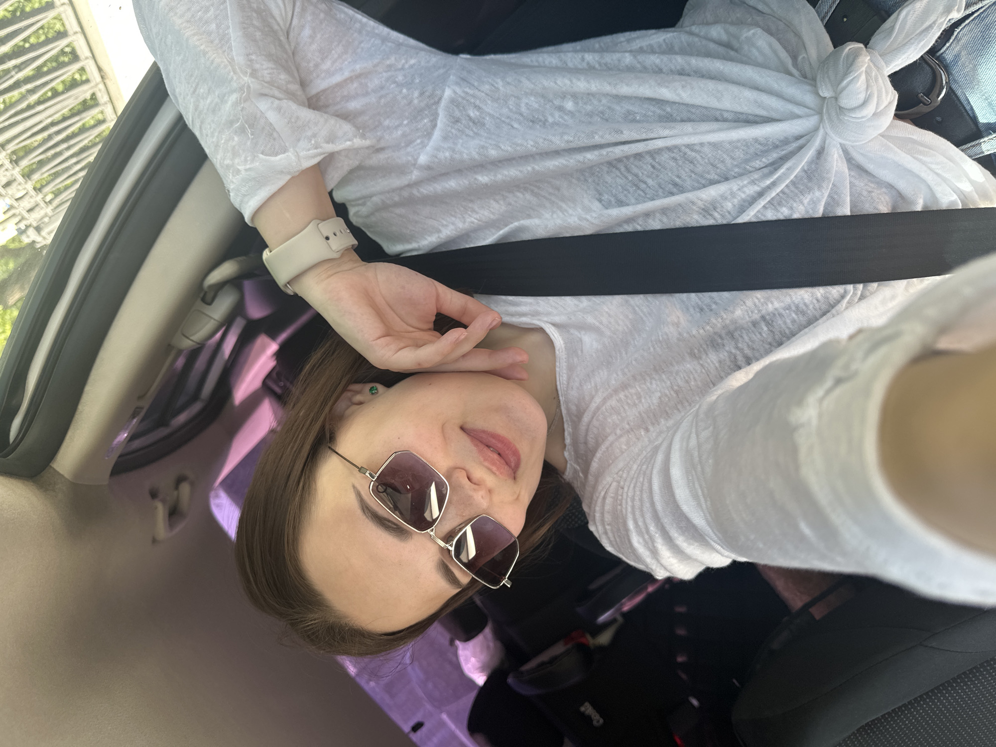 A selfie taken inside a car, possibly in Perm, Russia, shows a woman wearing sunglasses and a white shirt knotted at the waist, secured by a seatbelt. She is in the driver's seat, and her watch is visible on her left wrist. Car interior elements are visible, along with a glimpse of greenery outside the window. The car seat and child safety seat are visible.