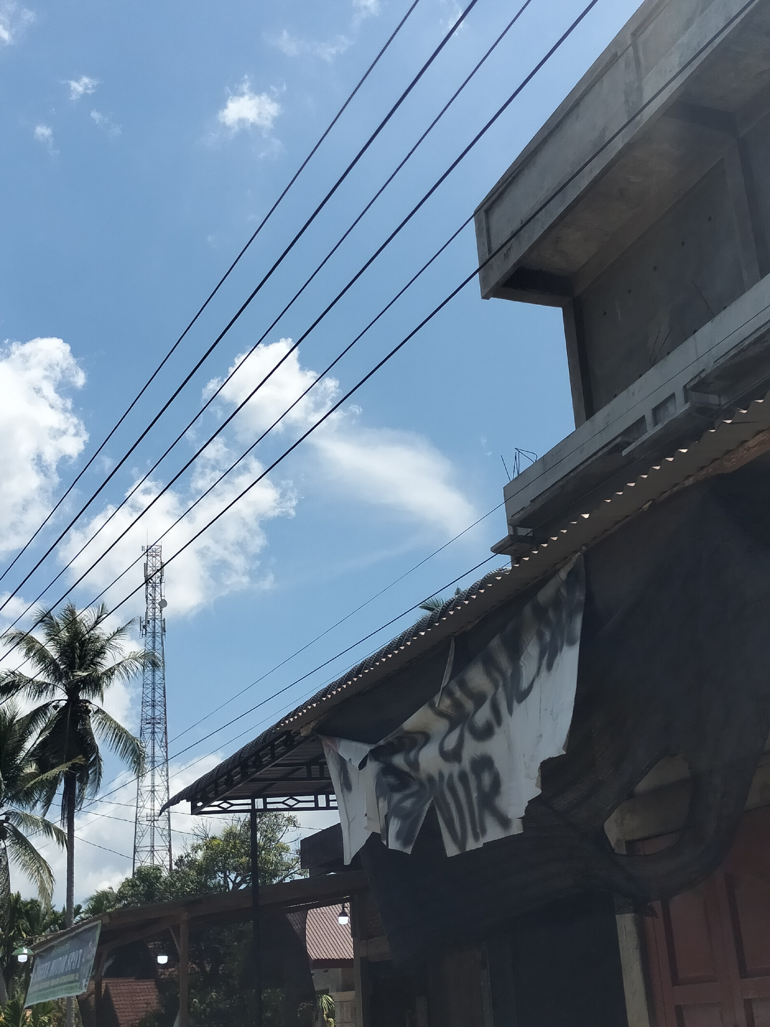 The image, taken in Matangpudeng, Indonesia, presents a low-angle view featuring a mix of natural and man-made elements. A partially obscured building dominates the right side of the frame, with unfinished concrete construction visible above the roofline. Below the concrete, a corrugated metal roof extends, and a tattered white banner with indistinct graffiti hangs limply from the awning. Adjacent to the building, an old tire is visibly mounted. Power lines stretch across the sky from the upper left to the upper right corner of the frame. Beyond the building, a tall communication tower is visible, partially obscured by the foliage of palm trees on the left side of the frame. The sky is a bright blue with scattered white clouds. Other buildings with roofs of brown color are also seen in the image.