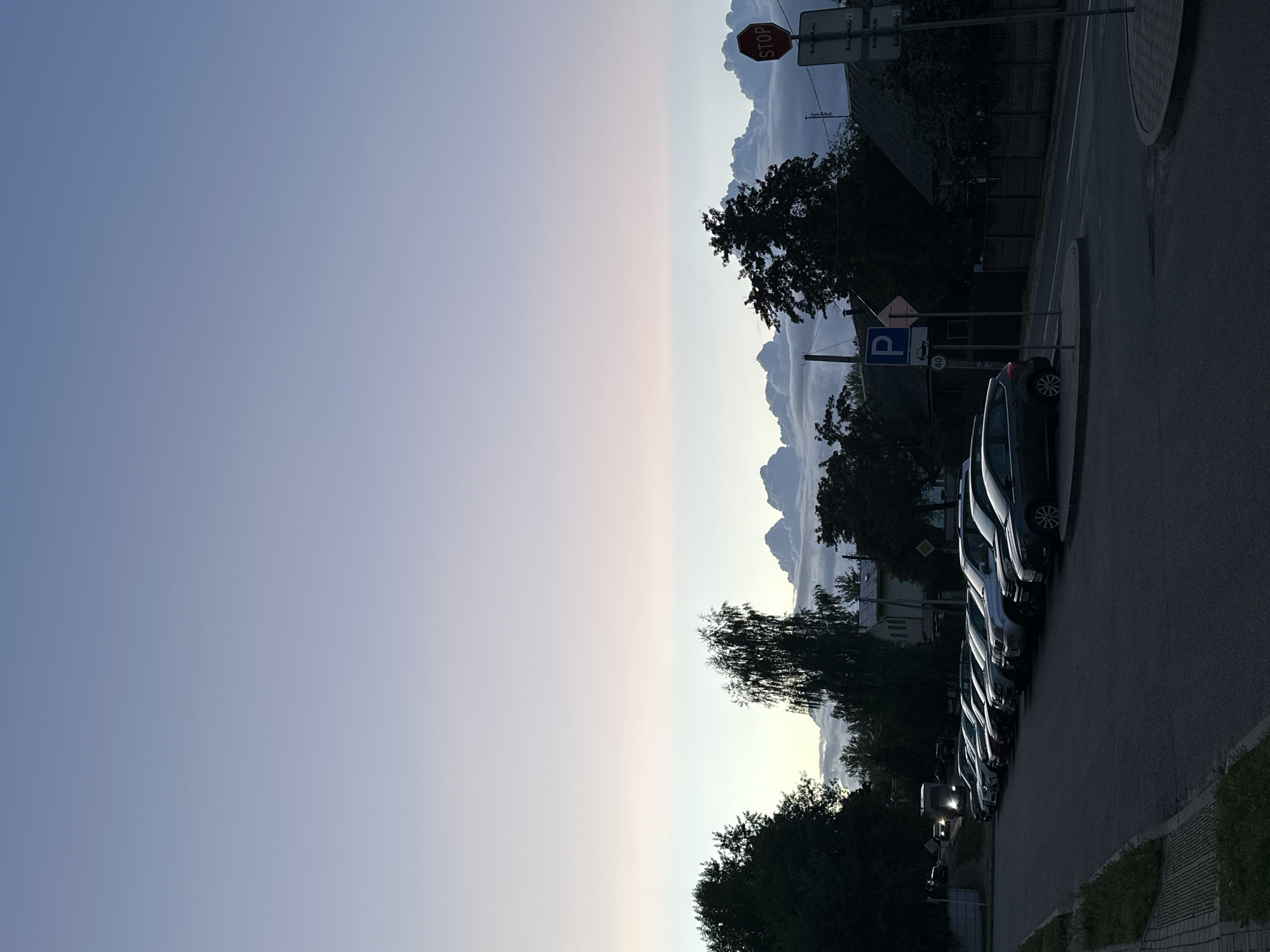 The image depicts a street scene in Minsk, Belarus, during twilight. A row of parked cars lines the asphalt road. In the background, trees and houses are visible. A stop sign and a parking sign are mounted on posts along the roadside. The sky above is a gradient of pale blue and pink, with a band of fluffy clouds low on the horizon. The overall lighting suggests it is either early morning or late evening.