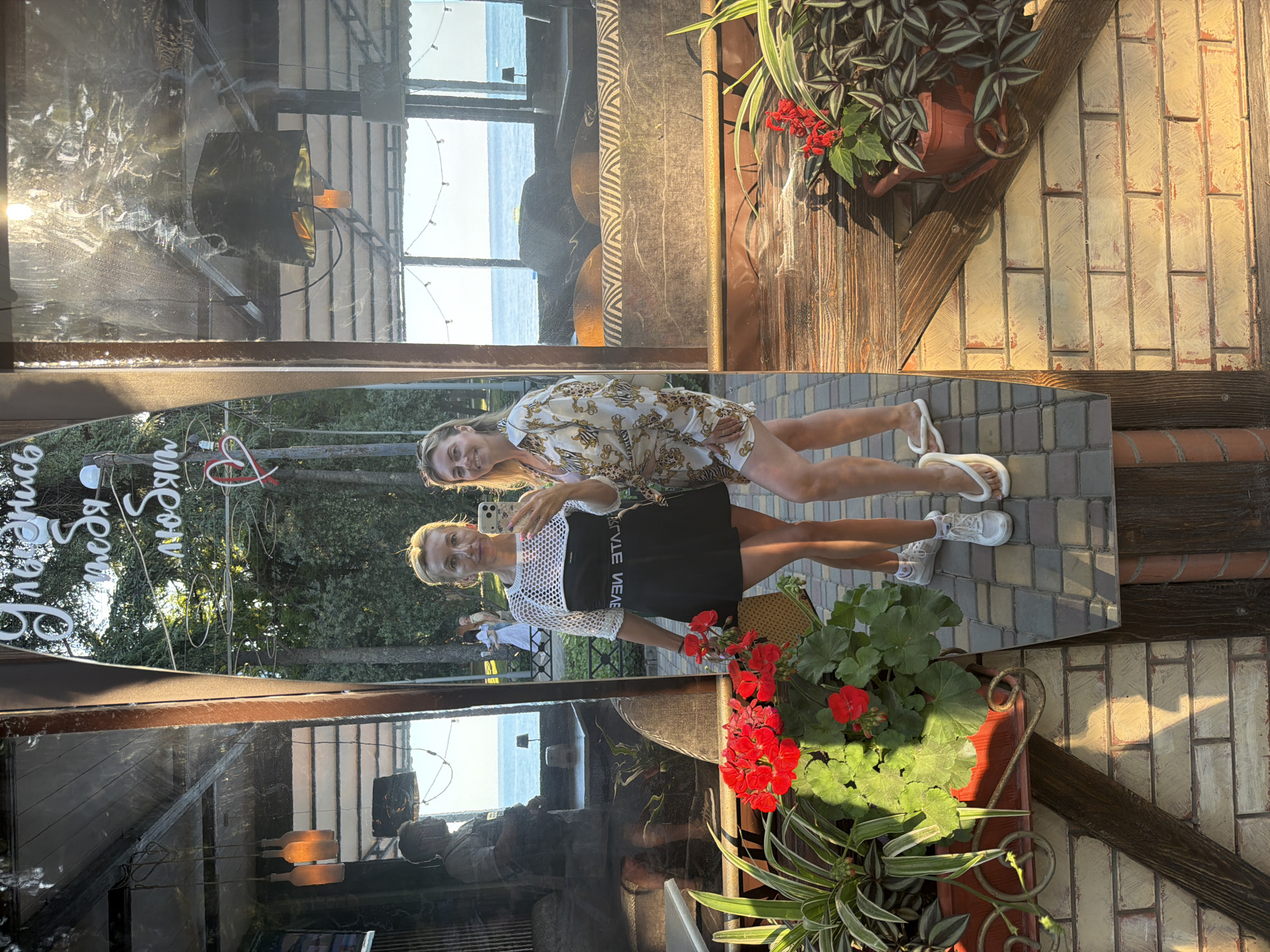 Two women are posing for a mirror selfie on a sunny day. The woman on the left is wearing a white crocheted top over a black dress, and white sneakers. She is holding a bouquet of red flowers. The woman on the right is wearing a white and gold patterned shirt and shorts, and white flip-flops. They are standing on a cobblestone patio in front of a building with a mirrored facade. In the background, there is a view of the Black Sea and trees. The mirrored facade reflects the interior of a cafe with hanging lamps and seating, as well as other patrons. The signage on the mirror is in Cyrillic script. Based on the context and prominent signage mentioning Alushta and Crimea, the location is Alushta, Ukraine.