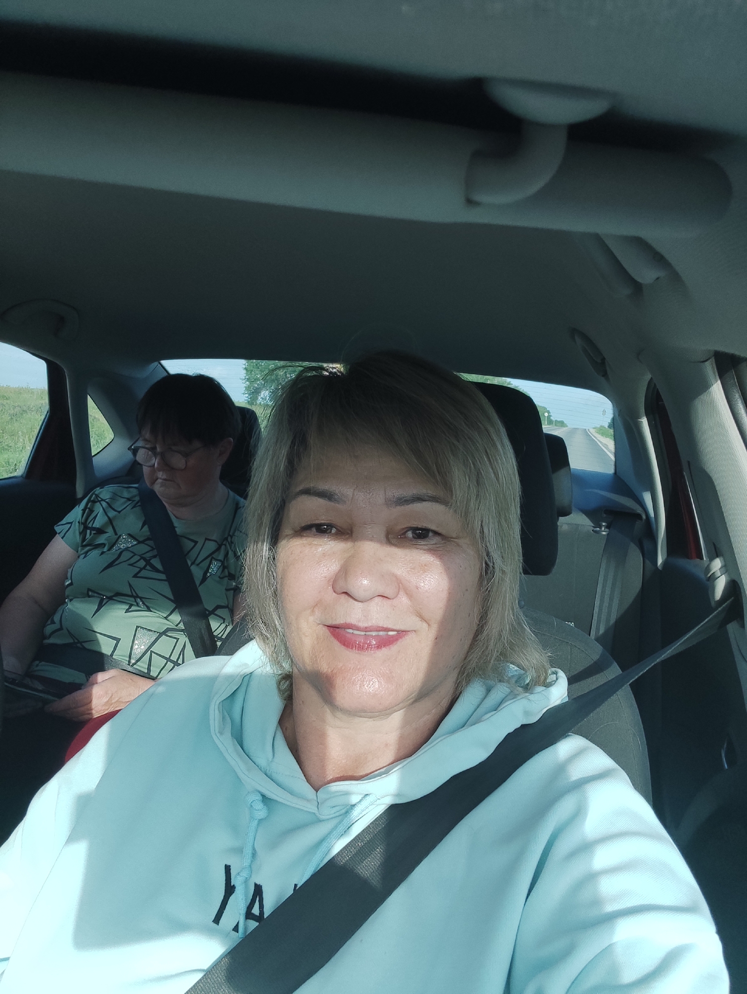 Two women are seated in the passenger and back seats of a car. The woman in the foreground is taking a selfie, smiling with her mouth slightly open. She has blonde hair and is wearing a light blue hoodie with the word "YARA" in black lettering. A gray seatbelt is across her chest. Behind her, the woman in the back seat, wearing glasses and a green patterned shirt, looks down and to her left. She also has a gray seatbelt across her chest. The car interior is visible, with the dashboard, ceiling, and seats. Outside the car windows, a rural landscape with green fields and a road stretching into the distance is visible, suggesting the car is in motion on a sunny day. The setting is the city of Staroilikovo, Russia.