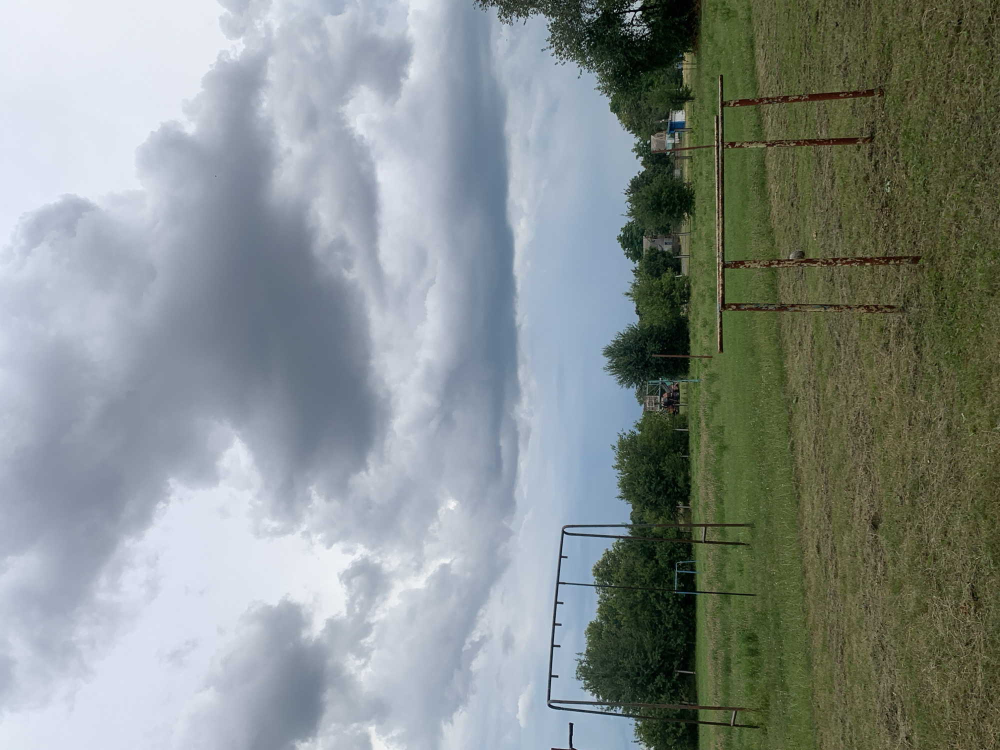 This image depicts an outdoor scene with a cloudy sky, featuring a grassy field with two sets of rusty metal exercise equipment. In the foreground, a pair of parallel bars stands on four legs. Further back, a horizontal bar with multiple hanging points is supported by two uprights. Trees and some small buildings are visible in the background. The scene appears to be located in a rural or semi-rural area, potentially a public space or an abandoned sports ground. The weather appears overcast, suggesting a potentially damp or cool day.