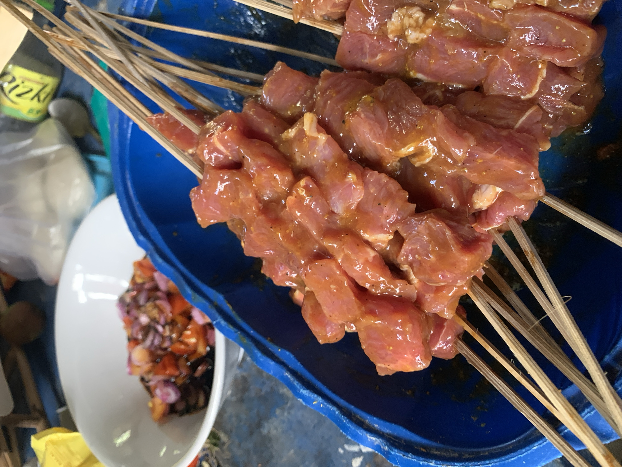 The image shows marinated meat skewers in a blue container and a bowl of chopped ingredients, likely a condiment or sauce. It was taken in Karangkemiri, Indonesia. The skewers appear to be wooden. The meat is raw and reddish in color. The chopped ingredients include red onion and tomato. There are bottles and miscellaneous objects visible in the background.