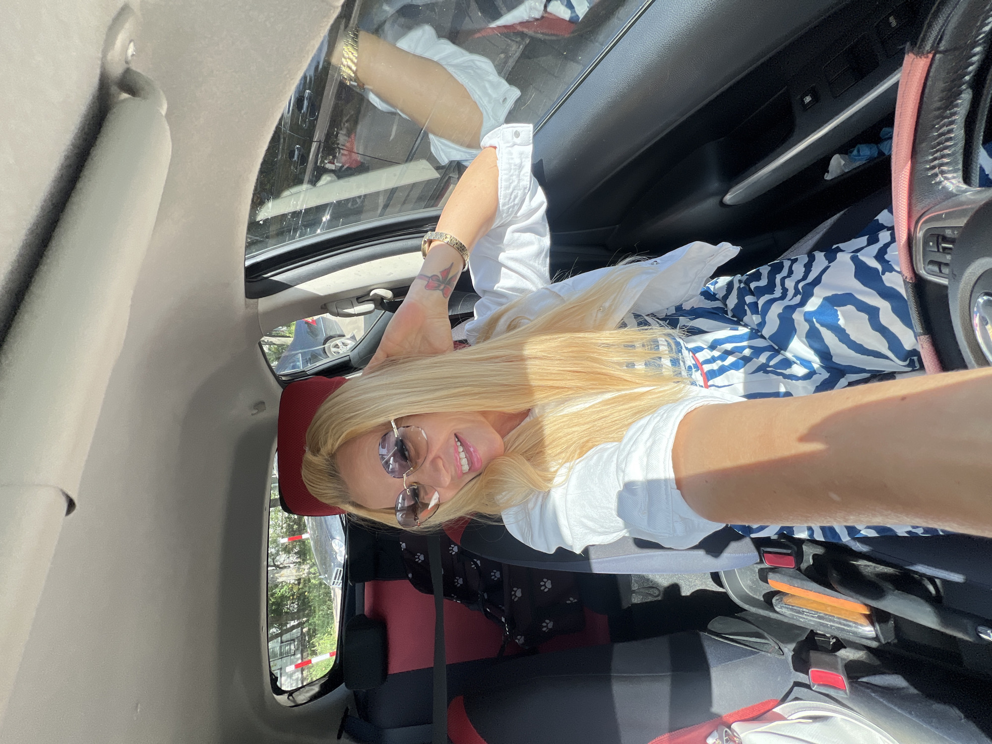 A blonde woman is sitting in the driver's seat of a car and taking a selfie. She is wearing heart-shaped sunglasses, a white shirt, and a blue and white patterned dress. She is smiling and her hair is blonde and wavy. The car is parked on a street in Białystok, Poland, with trees and other cars visible in the background. The interior of the car has red and black seats, and there is a steering wheel in the foreground. The sunlight is casting shadows on the woman and the car interior.