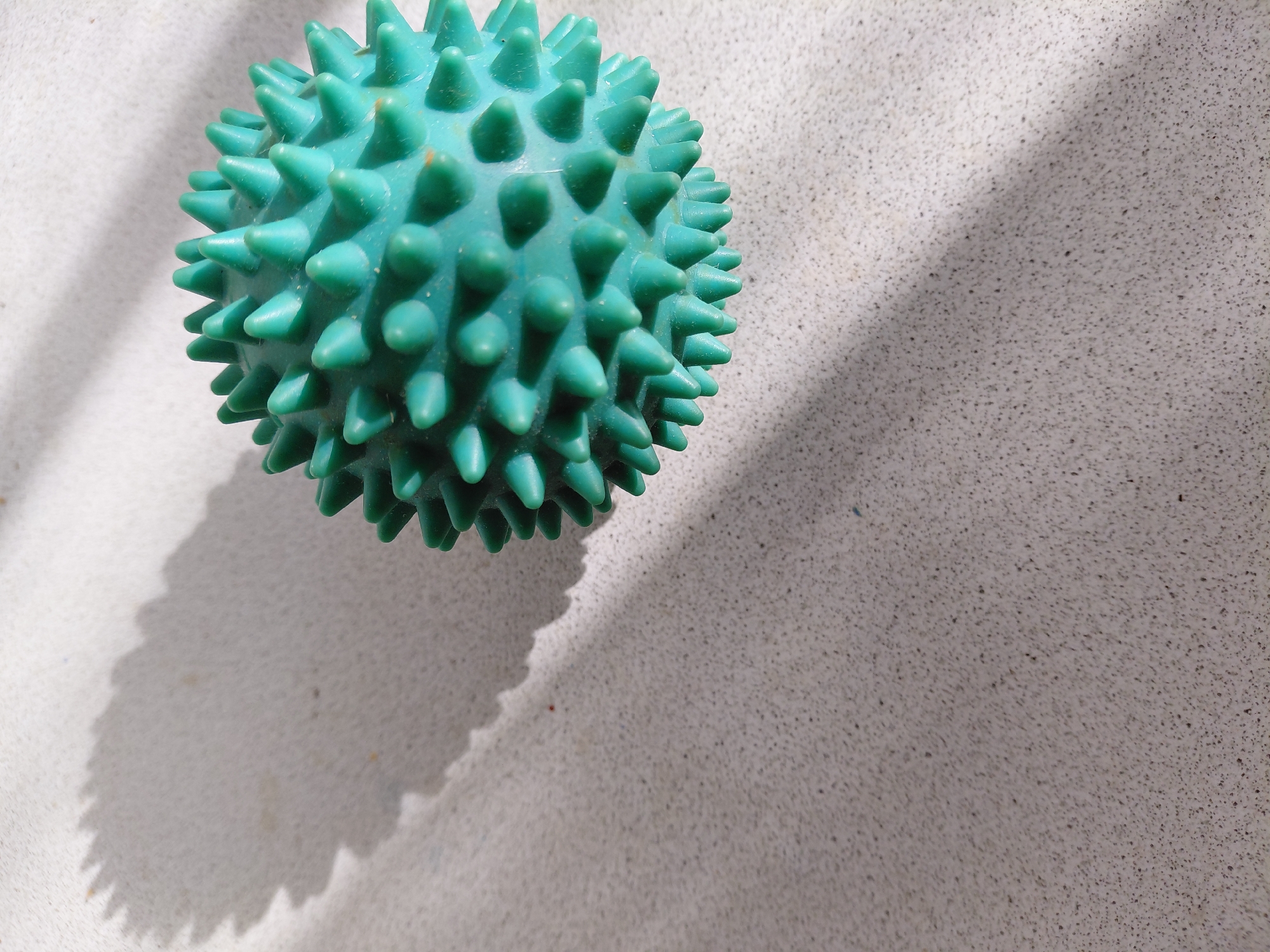 A teal-colored, spiked massage ball rests on a textured, light-colored surface, possibly concrete or stone. The ball is positioned in the upper right quadrant of the image. A shadow, cast by an unseen object, drapes across the left side of the frame, creating a serrated pattern on the surface. The lighting suggests an outdoor setting, with distinct shadows indicating sunlight. The image is a close-up shot, focusing on the texture of the surface and the details of the massage ball. There are no people or animals visible in the frame. The location of Zhukovsky, Russia, is not visually discernible from the image itself.