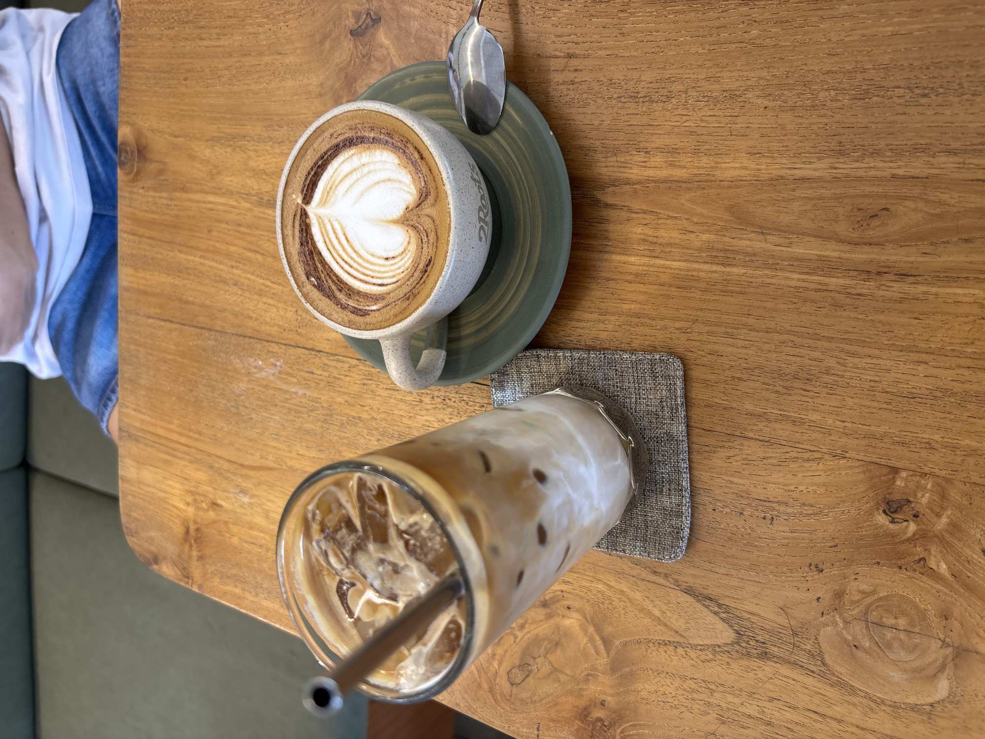 In Kuta Selatan, Indonesia, two coffee drinks are presented on a wooden table. To the left, a tall glass filled with iced coffee and a metal straw rests on a grey coaster. To the right, a ceramic cup with latte art in the shape of a heart sits on a green saucer, accompanied by a silver spoon. A person wearing a white t-shirt and denim shorts is partially visible in the background, seated on a green couch.
