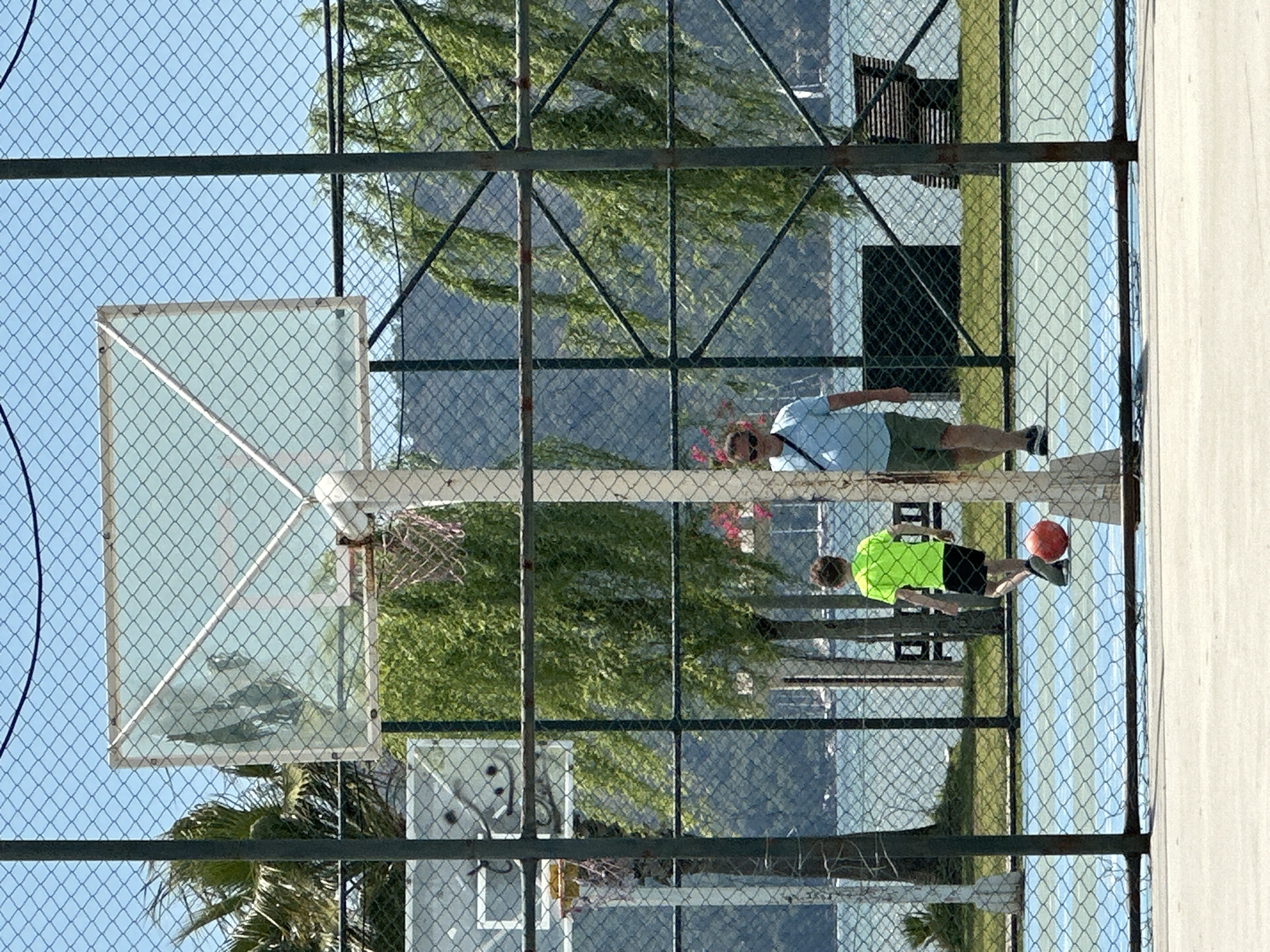 A basketball court in Fethiye, Turkey, is viewed through a chain-link fence. A basketball hoop and backboard stand at the left edge of the court. Two people are on the court: an adult male wearing a light blue shirt and khaki shorts and a youth wearing a bright green shirt and black shorts. The youth is holding a basketball. The court is made of light gray concrete. In the background, there are green trees and a body of water.