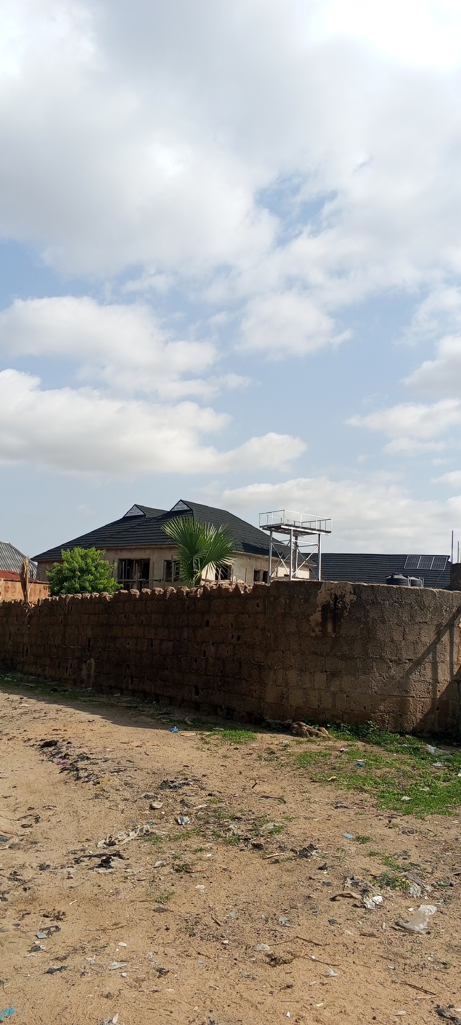 The image depicts a residential property in Jalingo, Nigeria, viewed from a dirt road. A tall, roughly constructed stone wall, approximately two meters high, dominates the foreground and runs horizontally across the frame. Beyond the wall, a house with a dark, multi-peaked roof is visible. To the right of the house, a metal water tower with a platform at the top stands, and further to the right, solar panels are installed on a roof. The ground in front of the wall is unpaved and littered with debris, including plastic bags, wrappers, and other trash, with sparse green vegetation growing in patches. The sky above is partly cloudy with large, white cumulus clouds against a light blue sky. No people are present in the image.
