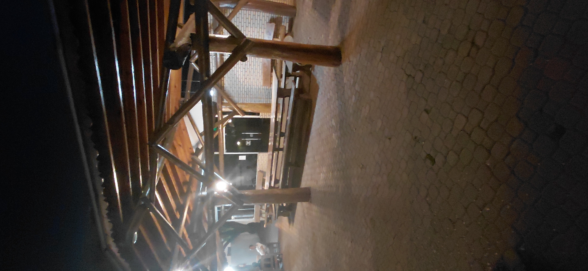The image shows an outdoor seating area under a wooden-framed roof at night. The area is paved with interlocking brick pavers. Several long wooden picnic tables with attached benches are arranged throughout the space. In the background, there is a building with brick walls and double doors. Lights are visible under the roof, casting shadows on the ground. Two people are seated at a table on the left side of the frame, and another person is standing near the tables on the left. The scene appears to be a casual dining or gathering spot, potentially a restaurant or bar, located in Presidente Franco, Paraguay.
