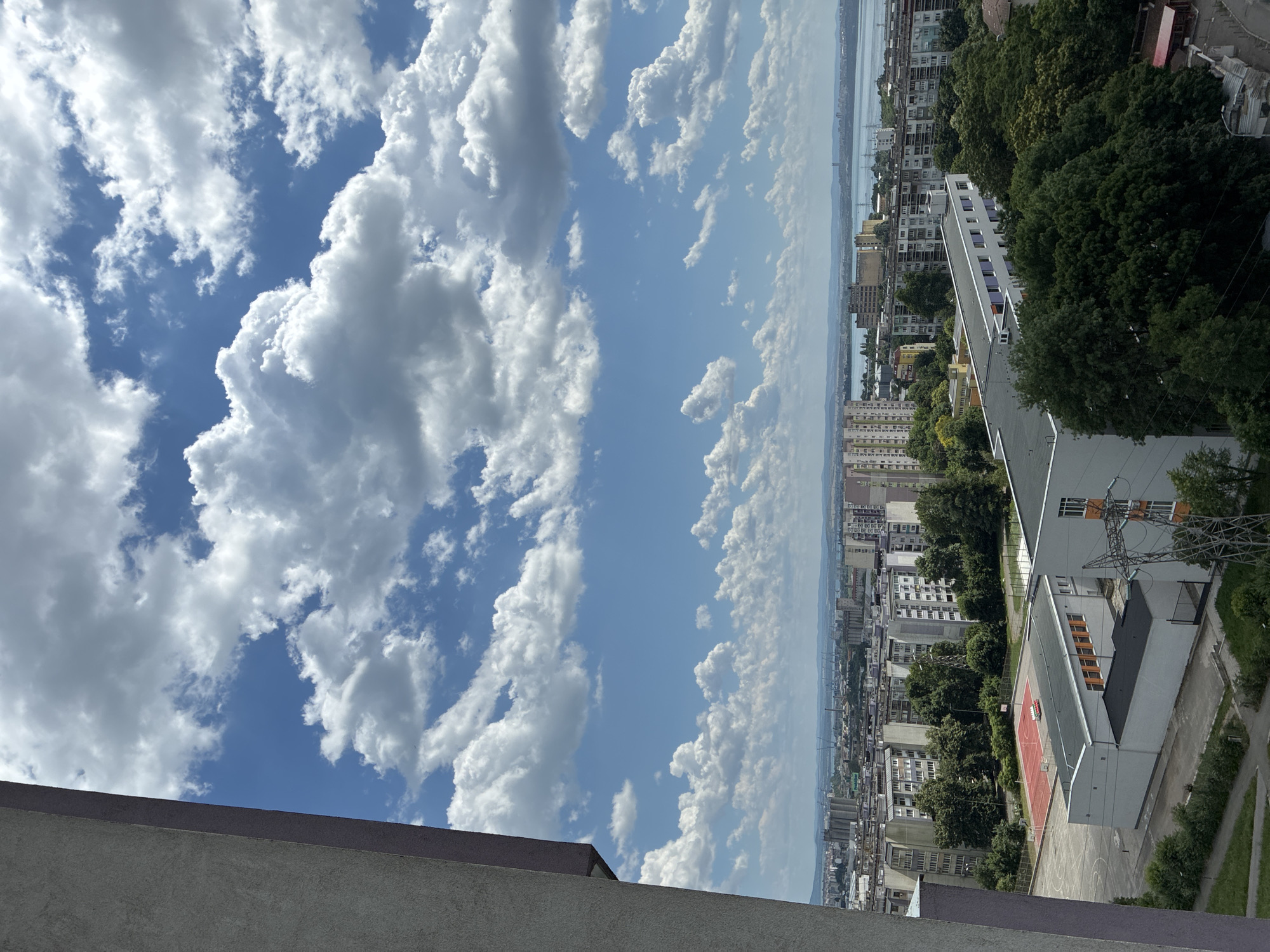 This is an aerial shot of a city with a body of water in the background. The sky is filled with cumulus clouds. In the foreground, on the left, is the side of a building. The middle ground is a cityscape with apartment buildings, trees, and a red basketball court. In the background, there are more buildings and a wide body of water, possibly a bay or a lake. The presence of apartment buildings and the overall urban landscape suggest a residential area within a city. The distant view of the water and some industrial structures could indicate proximity to a port or industrial zone. The time of day appears to be daytime, given the bright lighting and clear blue sky interspersed with clouds. Without specific landmarks or identifying features, pinpointing the exact city is challenging, but the architecture and urban planning could be characteristic of Eastern European cities. It is impossible to determine the specific location within Burgas, Bulgaria.