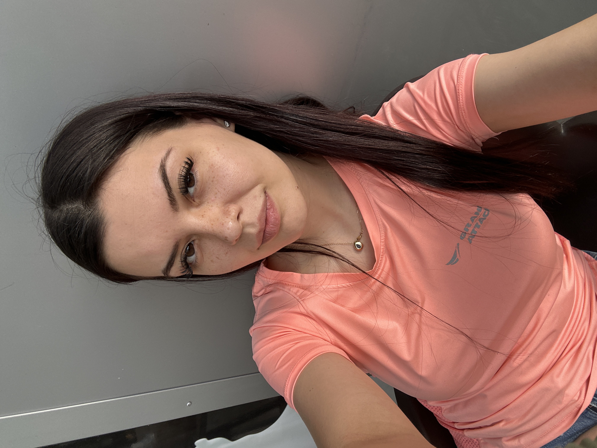 The image shows a young woman with dark hair and freckles, wearing a coral-colored t-shirt and a necklace with a circular pendant. She has on false eyelashes and appears to be taking a selfie against a plain gray background. The setting is indoors, likely a room or a studio, with a metallic surface visible on the left side of the frame. There are no discernible landmarks or elements that would suggest a specific location within Sliven, Bulgaria. The woman's pose is casual, and her expression is neutral, with her gaze directed towards the camera. The lighting is soft, highlighting her features and the texture of her clothing. The overall impression is that of a candid, personal photograph.