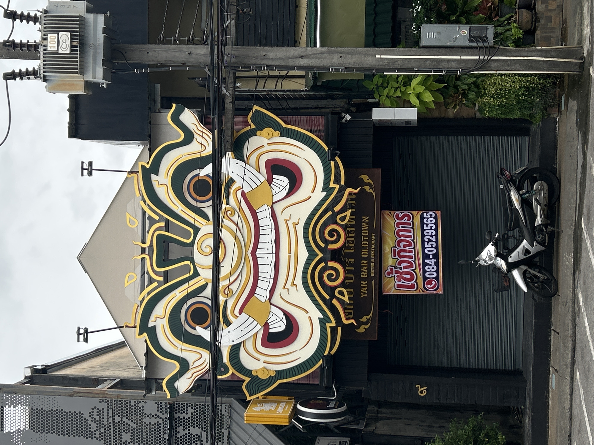 An exterior, eye-level, medium shot captures a large, ornate, masked facade above a closed storefront in Phuket City Municipality, Thailand. The mask, predominantly white with intricate gold and green detailing, features prominent yellow fangs, red lips, and large, round, blue and orange eyes. Below the mask, a dark brown sign reads "YAK BAR OLDTOWN BISTRO & RESTAURANT." A bright yellow sign with a red phone icon and Thai text, "เช็คคิวการ 084-0529565," is affixed to a grey roll-up shutter. To the left of the shutter, a gold Thai numeral is visible on a dark brick wall. A silver motorcycle is parked on the sidewalk in front of the shutter. Electrical infrastructure, including a transformer box and numerous wires, occupies the right side of the frame. Lush green foliage spills down from planters on the right. The sky is overcast.
