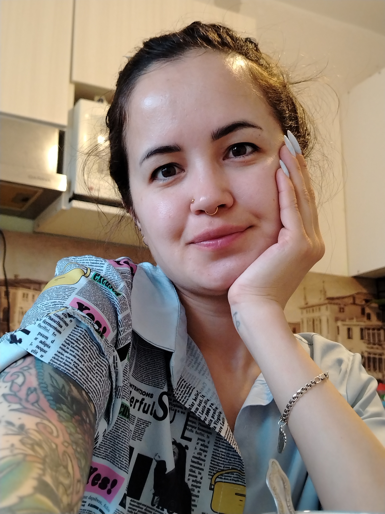 A woman with fair skin and dark hair pulled back in a loose bun poses for a selfie in an indoor setting. She has a nose ring and is resting her chin on her hand. Her fingernails are long and white. She wears a shirt with a newspaper print pattern and a bracelet on her left wrist. Her left arm has a full-sleeve tattoo. The background includes kitchen cabinets, an exhaust hood, and a print depicting a European cityscape. The city in the print is not identified. The specific location in Noviy, Russia is not evident from the image.
