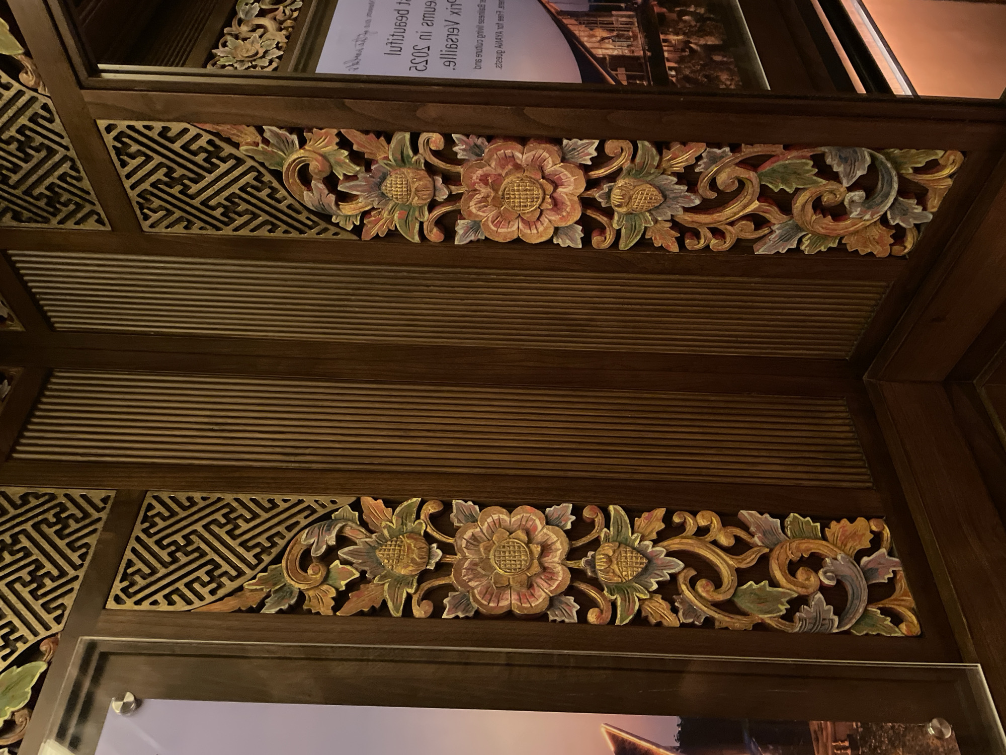 The image shows a close-up of ornate wooden carvings. The carvings feature floral motifs with central flowers flanked by stylized leaves and tendrils. The floral designs are colored with hints of orange, green, and purple. Interspersed within the floral patterns are sections of geometric latticework. The carvings are framed by dark wooden panels. The background consists of parallel wooden slats. The carvings are from a building or structure, most likely a museum, located in Kuta Selatan, Indonesia.
