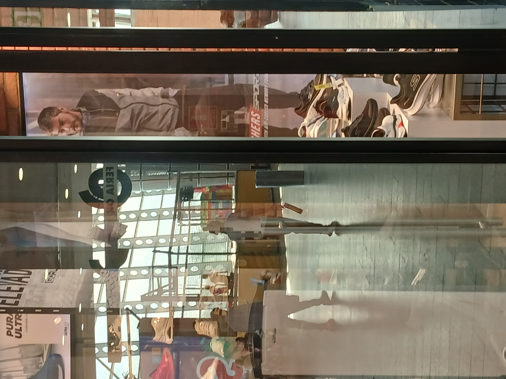 The image shows a storefront window of a Skechers shoe store. The window display includes a promotional banner featuring a male model wearing a jacket, next to the brand name "Skechers SPORT." There are also displays of Skechers sneakers. Reflections in the window show the interior of what appears to be a shopping mall. Based on location data, this is Ciudad Guayana, Venezuela.
