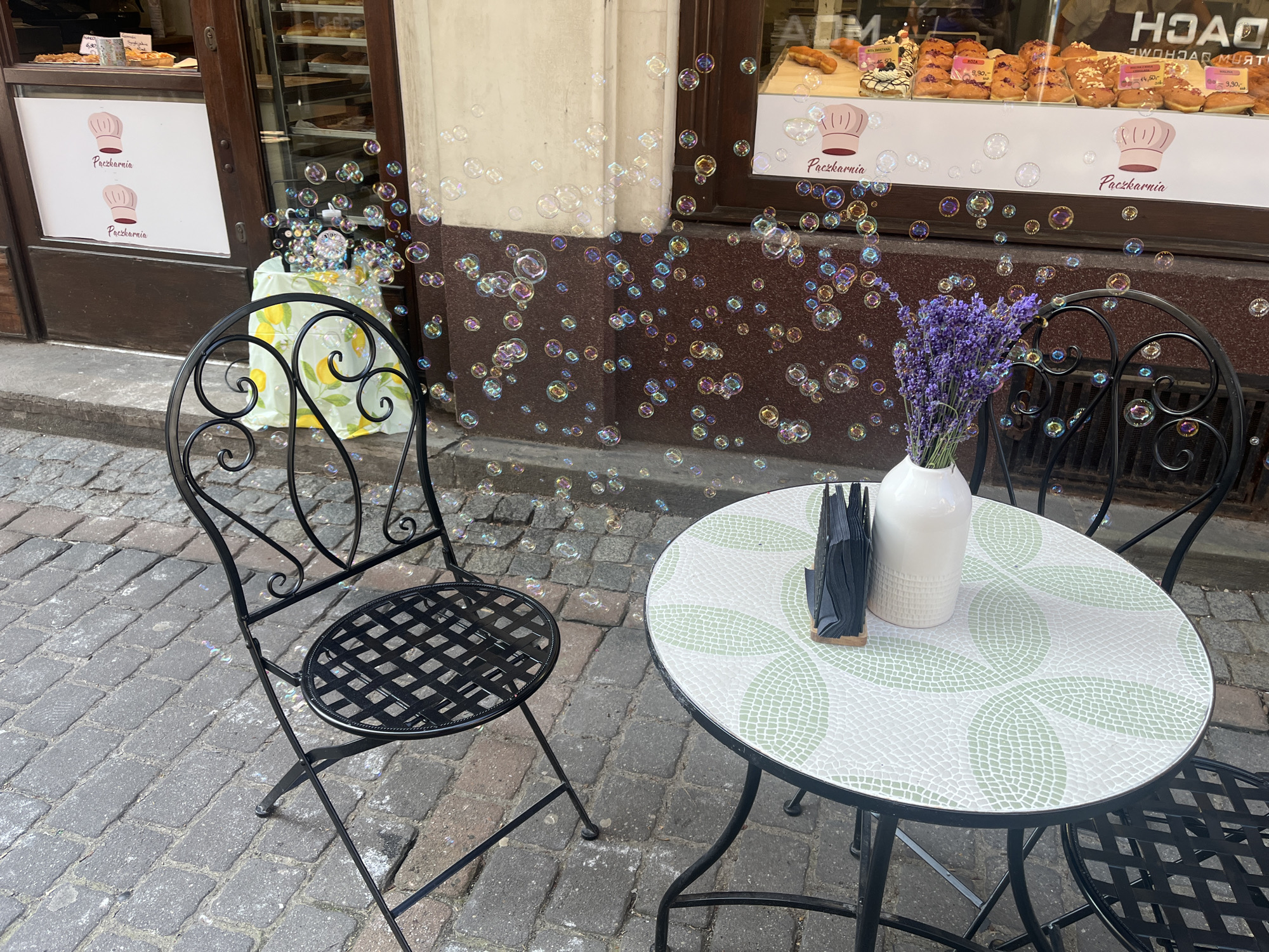 In a quaint European-style setting, a charming outdoor seating area is set for a delightful experience, surrounded by a whimsical display of iridescent soap bubbles. Two wrought-iron chairs and a matching table, adorned with a mosaic of green leaves on a white surface, are arranged on a cobblestone pavement. A white vase holds a bouquet of vibrant lavender, adding a touch of natural beauty to the scene. A napkin dispenser sits on the table, ready for use. Behind the seating area, the facade of a bakery, "Pączkarnia," is visible, showcasing a tempting array of pastries in its display window. The bakery's windows are decorated with two cartoonish chef hats, a playful touch that enhances the establishment's inviting ambiance. The atmosphere is serene and inviting, promising a relaxing moment amidst the bustling city.