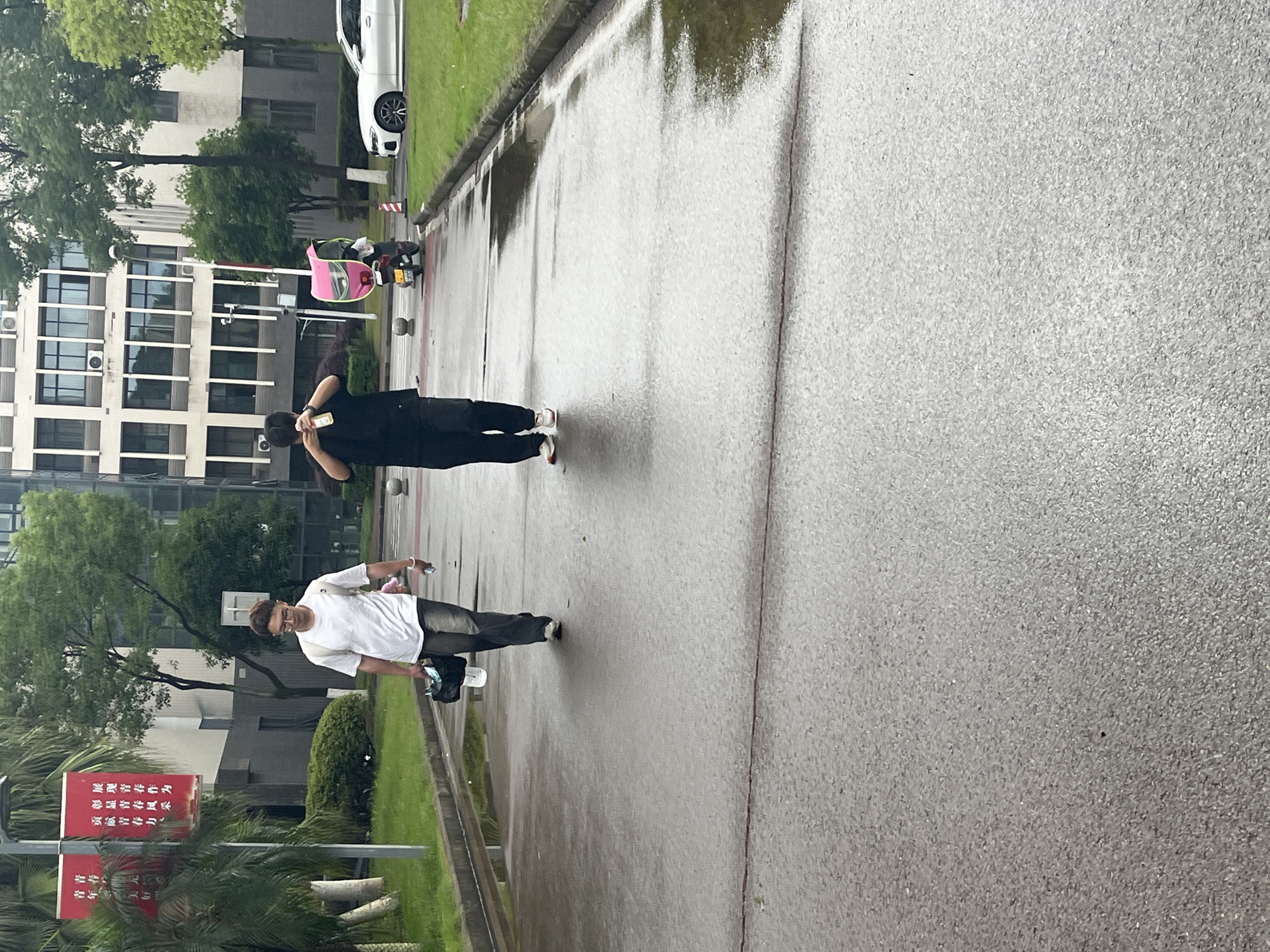 Two individuals are walking on a wet asphalt road outside a multi-story building. The person on the left, wearing a white t-shirt and dark pants, carries a black bag with a white bottle in one hand and holds a phone in the other. The person on the right, dressed in a black t-shirt and black pants, is holding a yellow bottle to their face. A pink delivery scooter is parked on the right side of the road, next to a white car. Red banners with Chinese text are visible on poles on the left side of the image. The environment appears to be a city street with green grass lining the road. The presence of the red banners and building architecture suggests the location is likely within China.