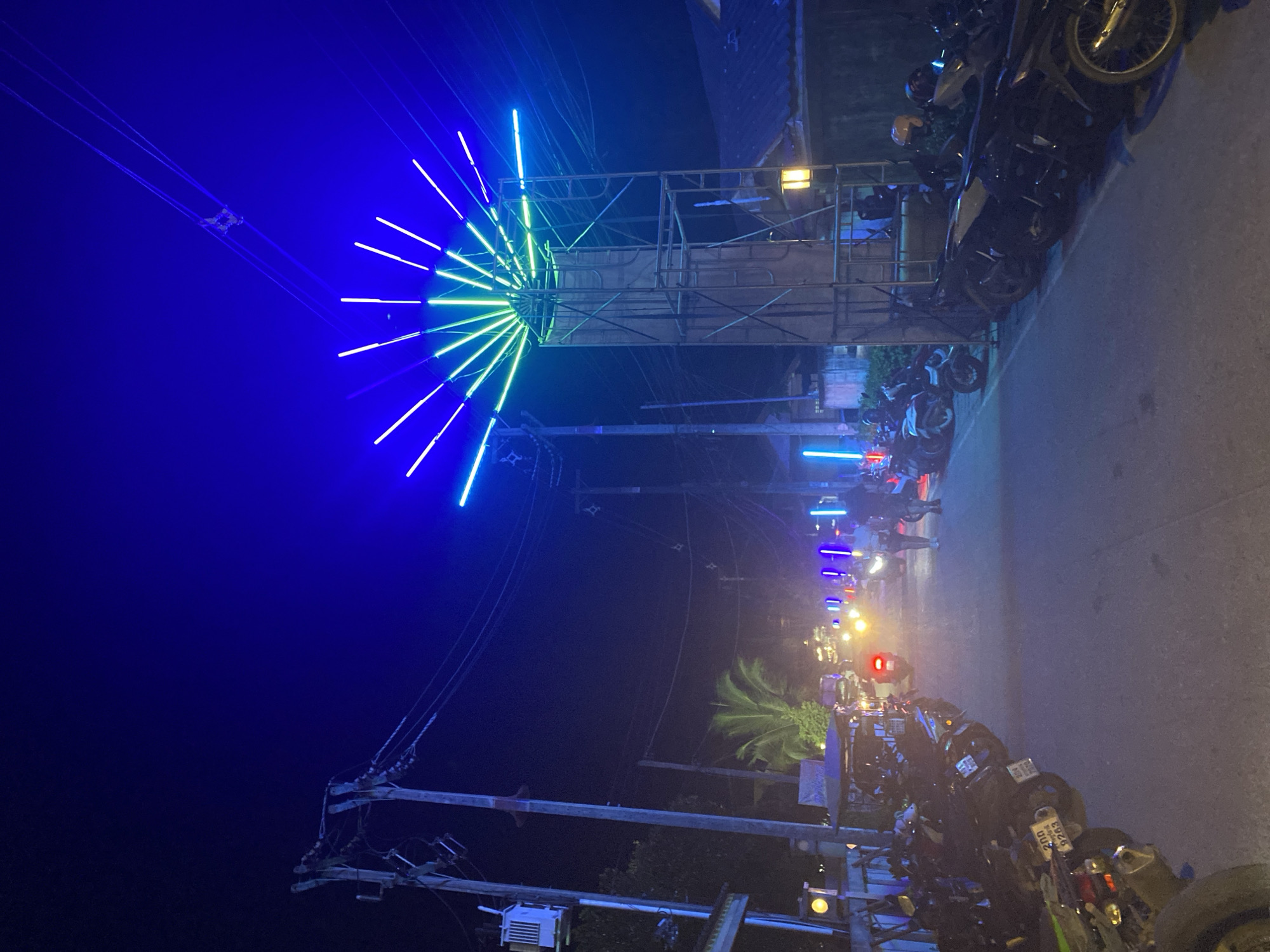 The image depicts a nighttime street scene in Baan Mae Nam, Thailand. The area is illuminated by a combination of streetlights, vehicle headlights, and decorative lighting installations. A large, radial light fixture, composed of blue and green neon lights, hangs from power lines above the street. Down the street, several vehicles are visible, including motorcycles. Motorcycles are parked along the side of the road. Overhead utility wires extend across the frame, connecting to wooden poles. The area appears to be a residential street or a busy thoroughfare, lined with parked motorcycles and possibly other vehicles.