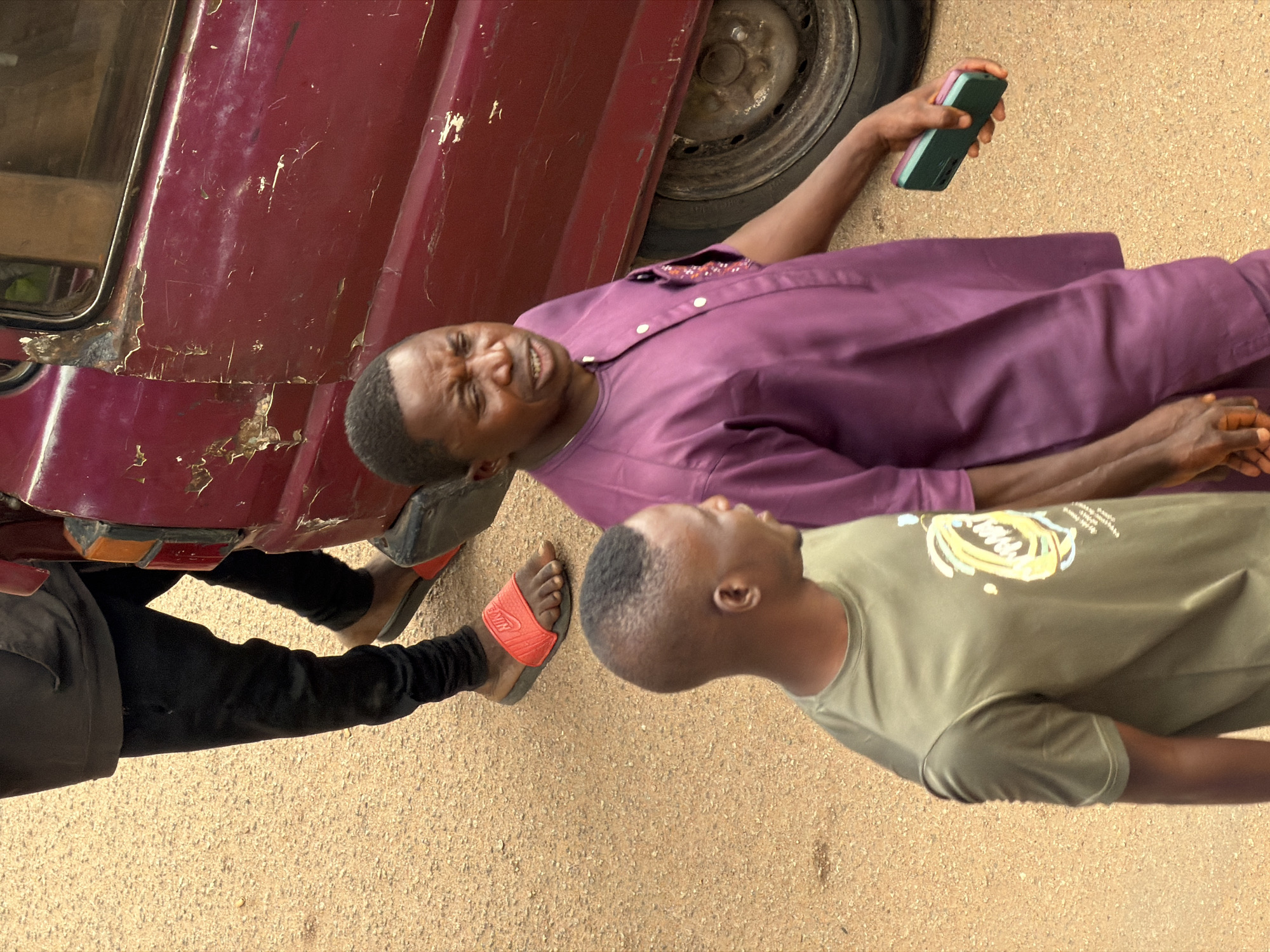 Two men are walking on a dirt road in Bukuru, Nigeria, next to a damaged burgundy colored vehicle. The man in the foreground is wearing a green t-shirt and appears to be looking back over his left shoulder. He has short hair. The second man is wearing a purple outfit and is holding a dark green and pink mobile phone in his right hand. He is looking forward and appears to be speaking or reacting to something. A third person's legs are visible on the left side of the image, wearing black pants and red Nike slides. The ground is a sandy, dirt surface. The vehicle appears to be old and has significant damage to its side, including rust and missing paint.