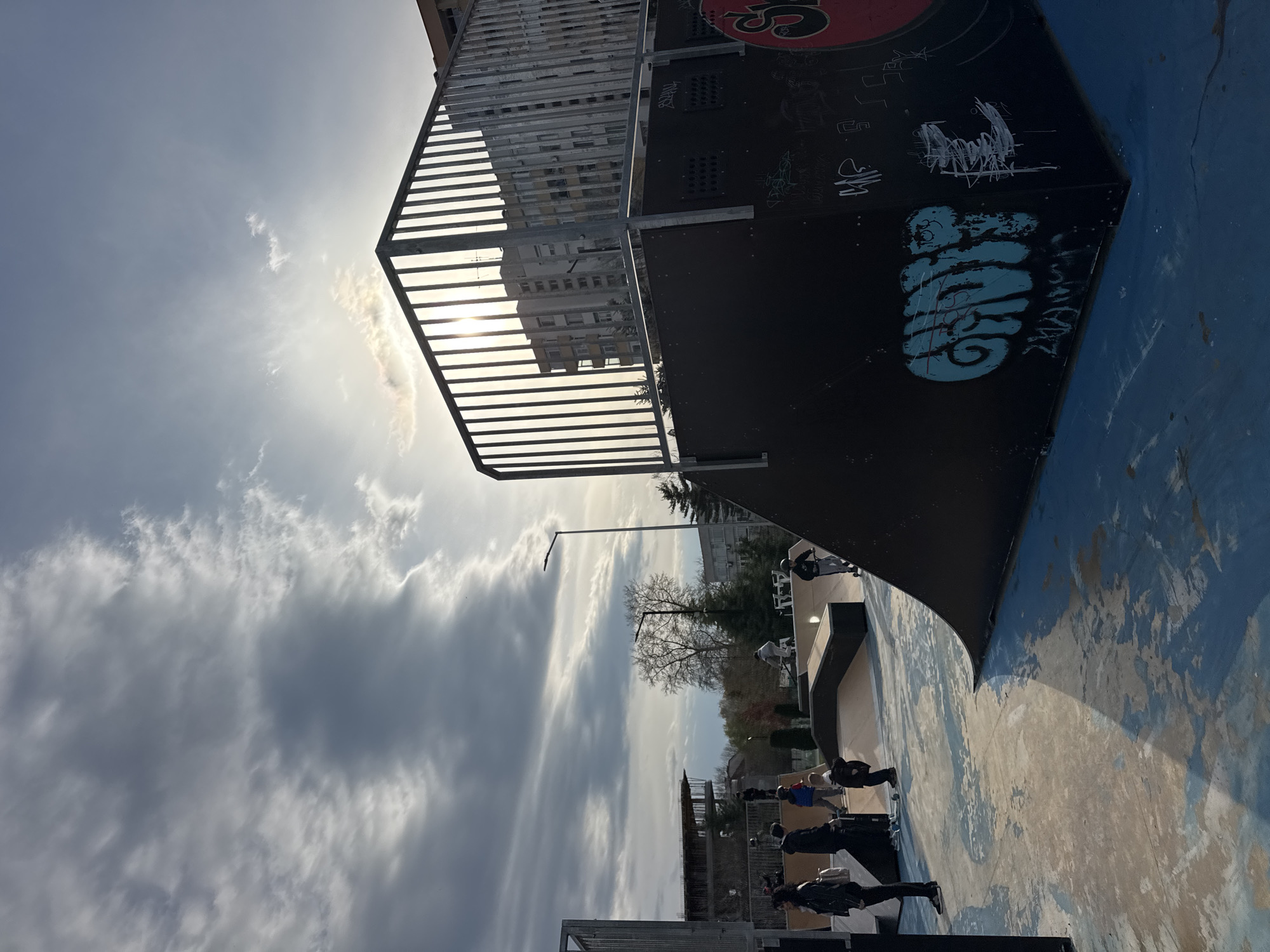 The image depicts a skate park in Blagoevgrad, Bulgaria. The scene includes a ramp with graffiti, a metal railing, and a concrete skating surface with blue paint. Several people are present, some walking and others seemingly engaged in skating or related activities. A building is visible in the background, partially obscured by the ramp. The sky is cloudy, with the sun breaking through in the upper left. A light pole stands in the mid-ground.