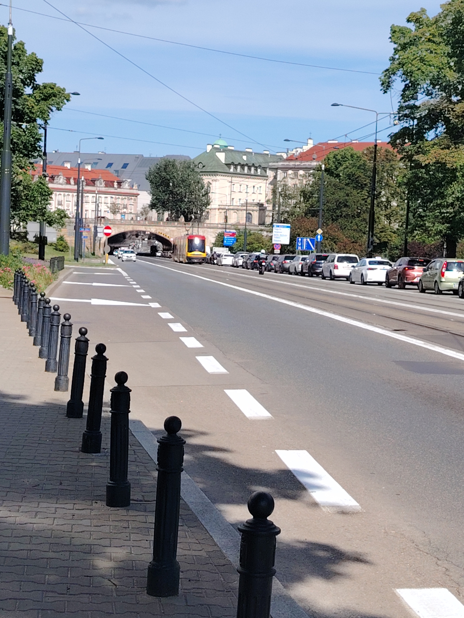 The image captures a street scene in Warsaw, Poland, likely in a central urban area, featuring a tram, cars, and pedestrians on a sunny day.
In the foreground, a paved sidewalk runs along the left side of the image, lined with black decorative bollards. A woman with dark hair is walking on the sidewalk. Adjacent to the sidewalk is a lane marked with white arrows indicating traffic direction, and dashed white lines separating it from the road.
The road itself is multi-laned, with tram tracks clearly visible. A yellow and red tram is traveling away from the camera on the tram tracks. Several cars are driving in the same direction, forming a queue.
In the background, buildings with red and white facades and green roofs are visible. An arched overpass crosses the street. The sky is blue with scattered clouds. The overall environment suggests a typical day in a busy Polish city.