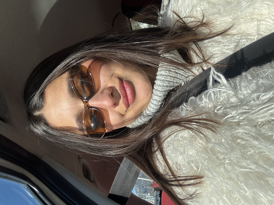 A female individual is depicted in a close-up, chest-up shot taken inside a vehicle in Zankovići, Montenegro. She has long, dark brown hair parted centrally and wears brown, oval-shaped sunglasses. Her attire includes a light grey ribbed turtleneck sweater and a white, shaggy-textured jacket. A black seatbelt is visible across her chest. Her lips are slightly parted, displaying a light pink color, and she presents a faint smile. Direct sunlight illuminates her face from the front-right, casting shadows. The vehicle's interior is partially visible, including a window frame on the upper left. Through the left window, a clear blue sky and distant, light-colored mountains are visible in the background.
