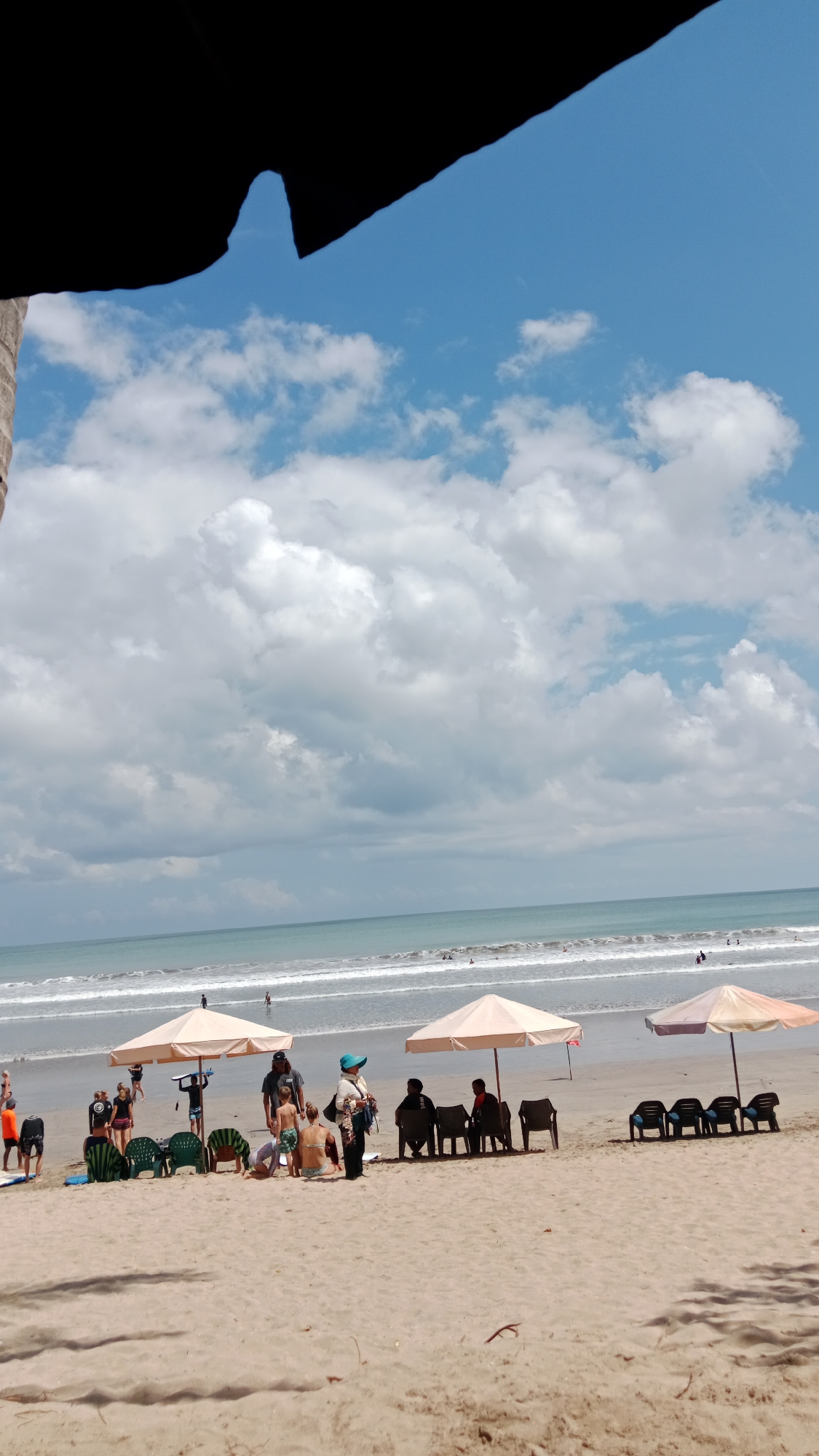 The image depicts a beach scene in Kuta, Indonesia, on a day with a partially cloudy sky. The foreground consists of a sandy beach with several groups of people and beach furniture. Umbrellas are set up to provide shade. In the middle ground, the ocean meets the shore with gentle waves. In the background, the horizon separates the sea from the sky, which is filled with a mixture of white clouds and patches of blue.
On the beach, there are at least four beach umbrellas, two of which are prominent and provide shade for groups of people. Under the leftmost umbrella, there are several empty green chairs and two people sitting. To the right of this, a group of people are gathered, including a person with a surfboard. Further to the right, under another umbrella, several individuals are seated on chairs. The rightmost umbrella also shades a row of chairs, some of which are occupied. People are dressed in casual beachwear, including swimsuits and shorts. Some individuals appear to be standing and talking, while others are seated. There are also a few people in the water, appearing to be swimming or enjoying the ocean. The sand is a light brown color, and there are some shadows cast on it, suggesting the sun is out. The overall atmosphere appears relaxed and typical of a beach day.