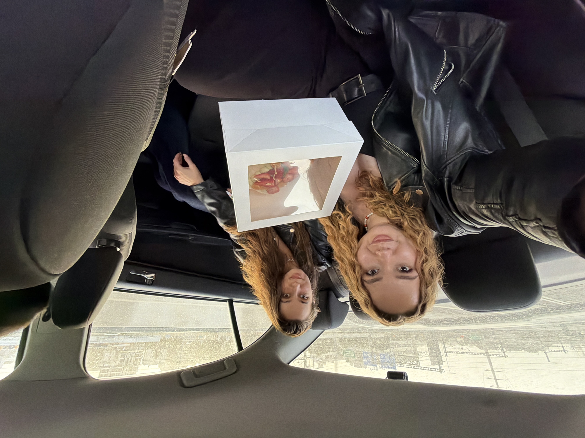 Two women sit in the back seat of a car, holding a bakery box. One woman wears a black leather jacket and a seatbelt. The car window shows a street scene in Tel Aviv, Israel.