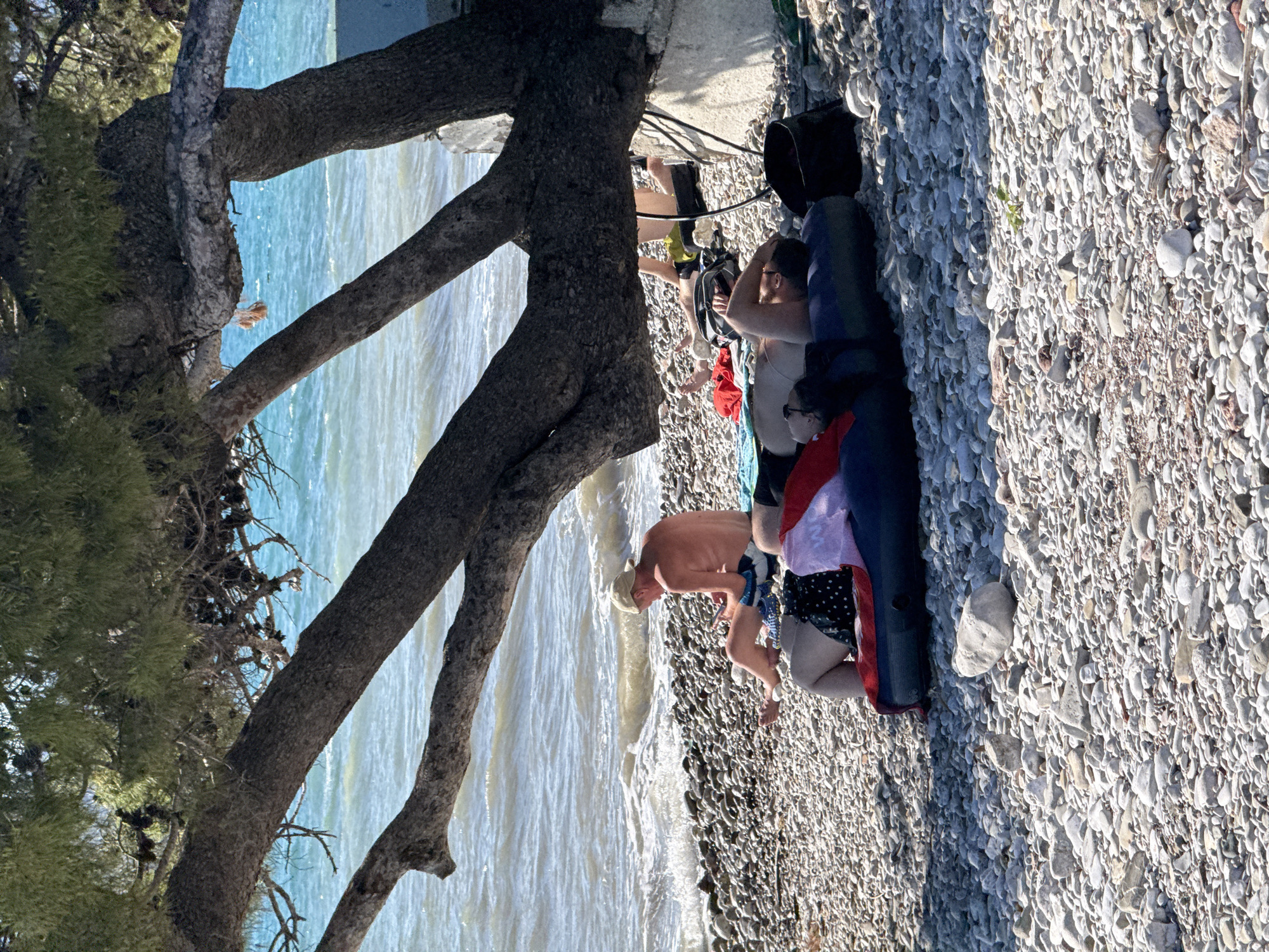 Two men are relaxing on a pebble beach in Bar, Montenegro, under the shade of a large tree. One man, wearing a light-colored baseball cap and patterned swim trunks, is sitting up and looking at his phone. He is positioned on a towel near the water's edge. The other man is lying down on an inflatable mattress, partially obscured by the tree. He is wearing a white tank top and appears to be resting his head on his arm. Several bags and a bucket are scattered around them on the beach. The sea is visible in the background, with gentle waves lapping the shore. The overall scene depicts a leisurely day at the beach.