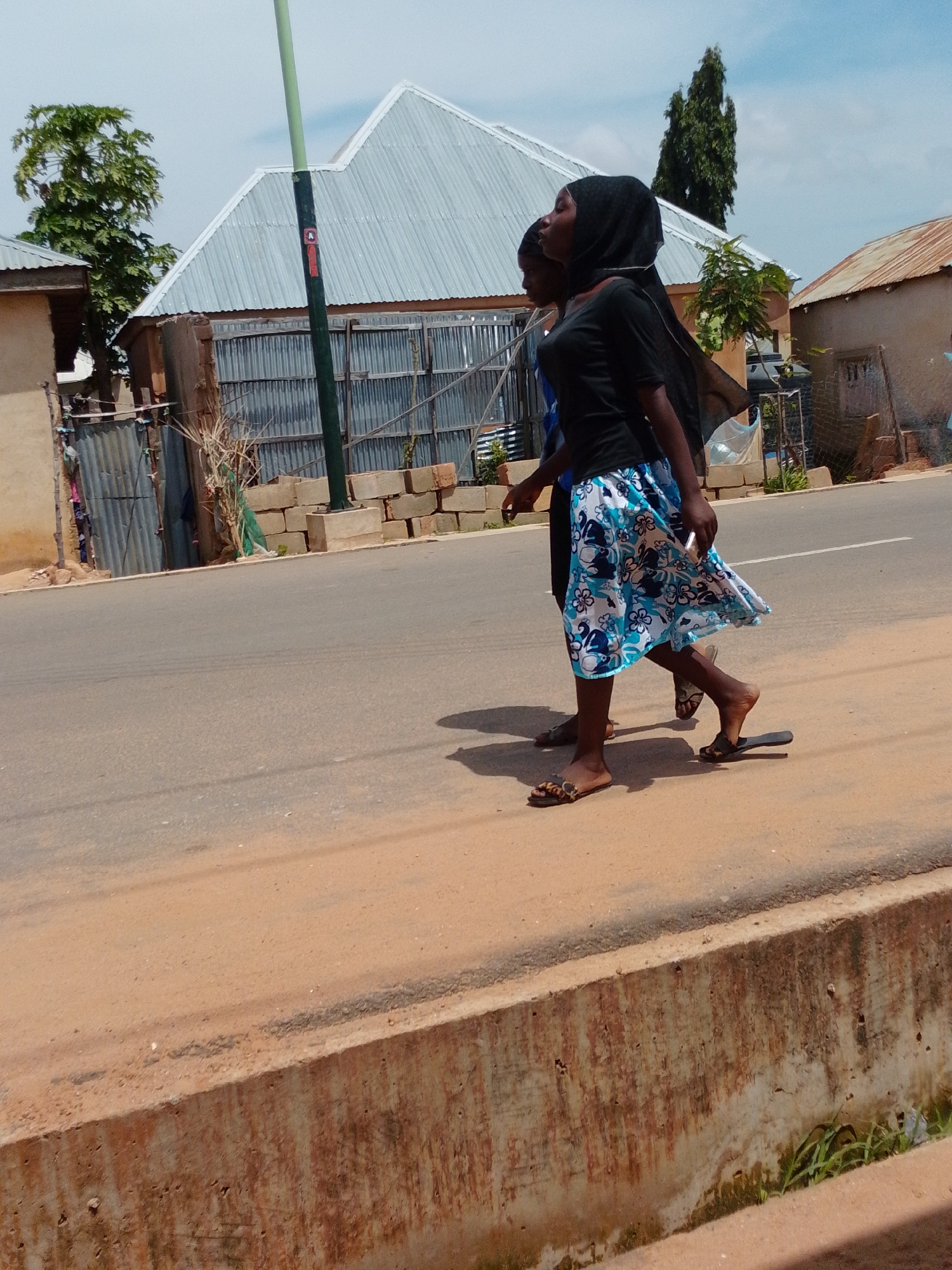 Two young women are walking on a paved road. The woman in the foreground is wearing a black t-shirt and a blue and white floral skirt, and a black hijab. She is looking to her left and holding a white object in her right hand. The second woman, walking behind and slightly to the left of the first, is wearing a blue shirt. The road is bordered by a dirt path and a concrete structure. In the background, there are several buildings with corrugated metal roofs, some trees, and a tall, narrow evergreen tree. The sky is partly cloudy.