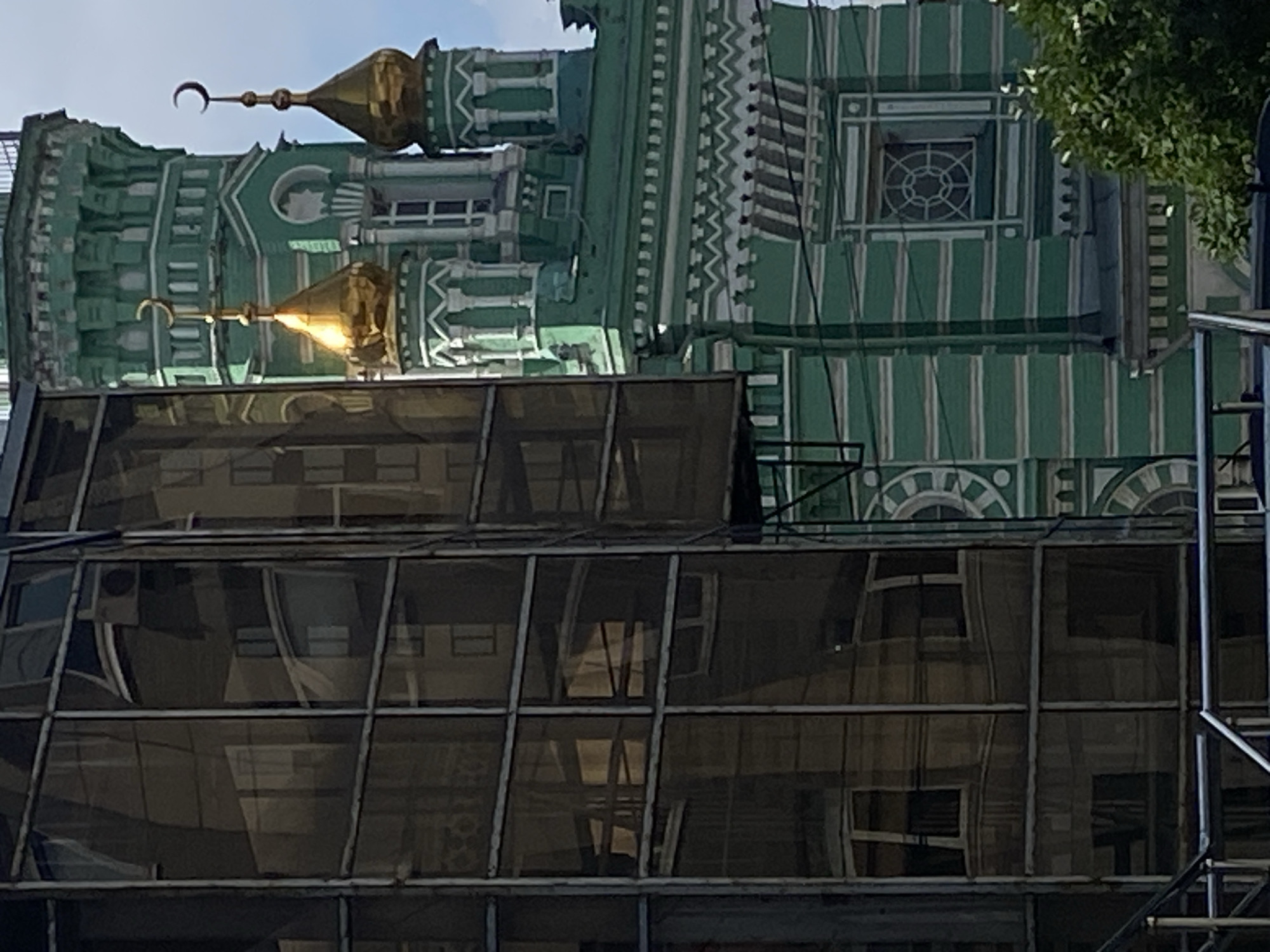 The image captures a close-up view of an ornate green building, likely a religious or historical structure, undergoing renovation or reconstruction. The building features decorative white trim and golden crescent-topped domes. In the foreground, scaffolding covered with a dark mesh material obscures the lower portion of the building, suggesting ongoing work. Reflections of windows and interior spaces are visible through the mesh, hinting at the building's internal structure. A glimpse of green foliage from a tree is visible in the upper right corner. The sky appears bright, indicating daytime. The overall scene suggests a construction site within an urban environment, possibly in Perm, Russia, given the location context. There is no visible text in the image.