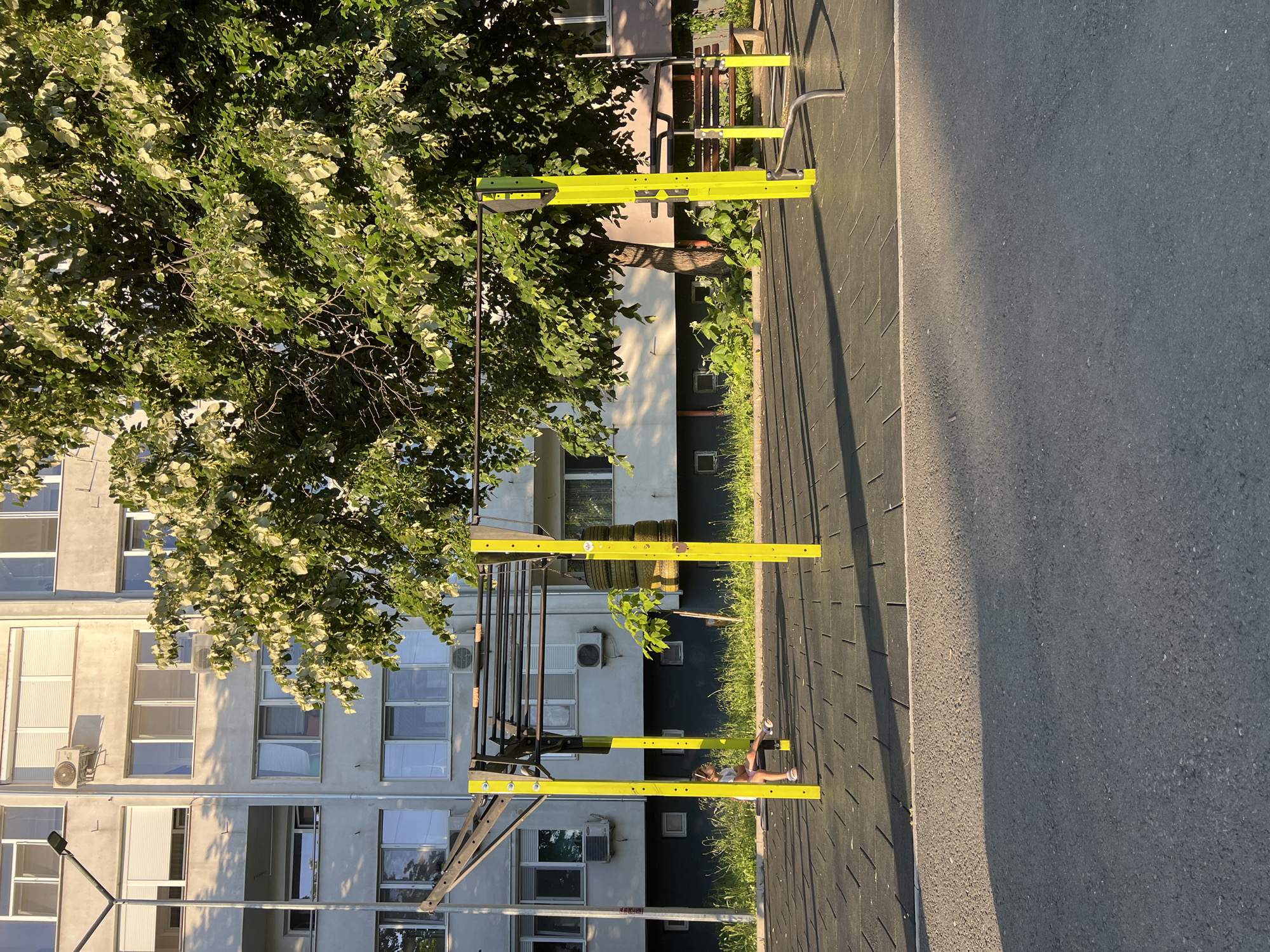 This is a photograph taken at an outdoor exercise area located in the city of Burgas, Bulgaria. The scene features an apartment building in the background, a large, leafy tree, and a piece of outdoor gym equipment constructed with bright yellow posts. A young girl is seated on a bench under the gym equipment. The ground is covered with a black rubber mat, and an asphalt surface is visible in the foreground. The sun is casting long shadows, indicating it is either early morning or late afternoon. The gym equipment includes horizontal bars and other elements designed for physical activity. The apartment building is multi-storied with visible windows and air conditioning units. Lush green vegetation grows around the base of the building and along the edge of the exercise area.