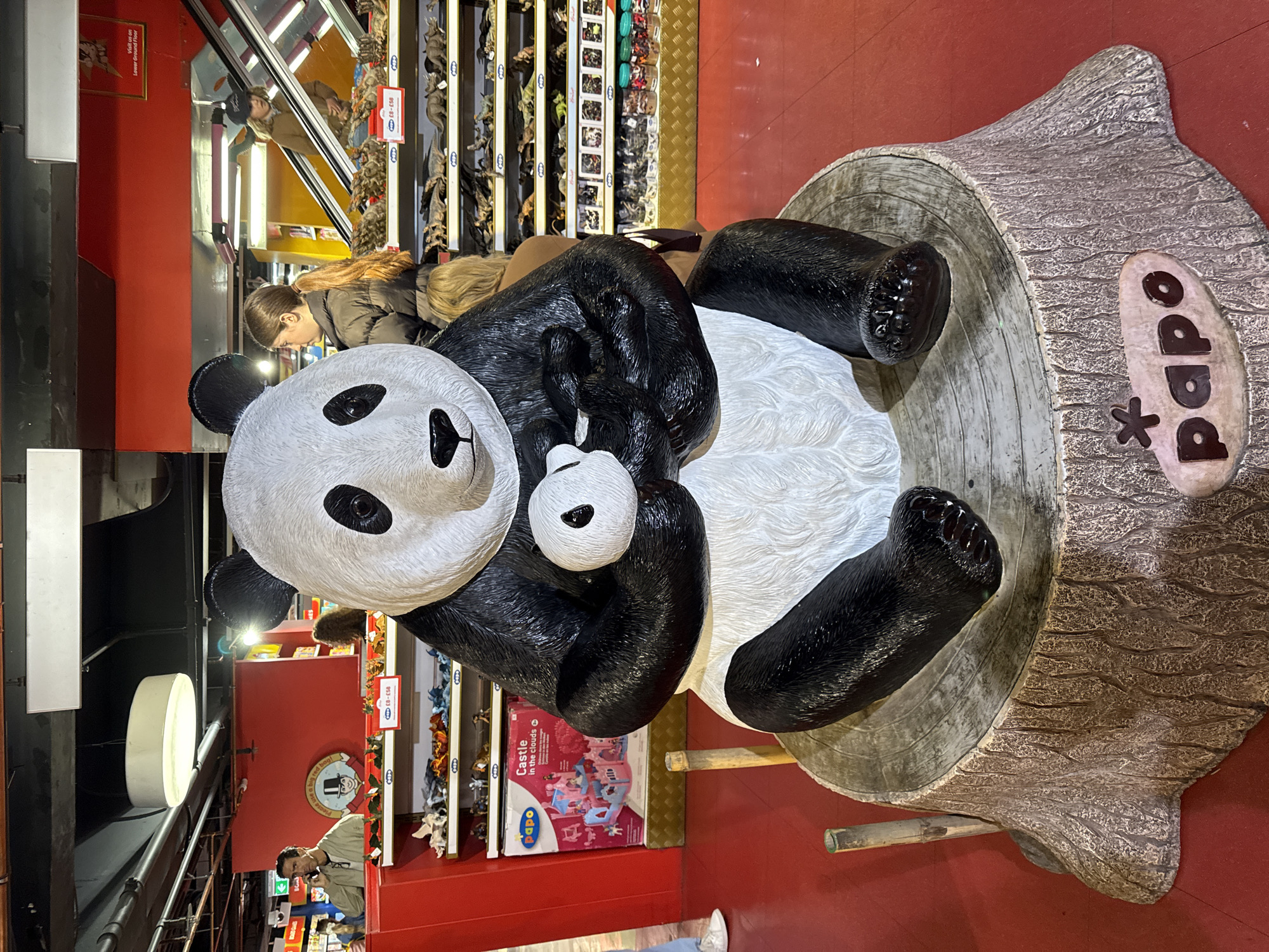 The image captures a scene inside Hamleys toy store in London, United Kingdom. The prominent subject is a large, decorative panda statue, seemingly made of plastic or resin, featuring a mother panda cradling a baby panda. The panda is positioned inside a faux tree stump with the "Papo" brand name on the side. To the left of the panda, a person in a light jacket appears to be talking on a phone, and an overhead circular light is visible. Behind the panda, an escalator can be seen with a woman looking down towards the panda. Adjacent to the panda are shelves filled with toy figurines and castle sets, and a sign advertising "Castle in the clouds" is noticeable. A sign reading "Visit us on Lower Ground Floor" is also visible. The floor is bright red, typical of the store's decor.