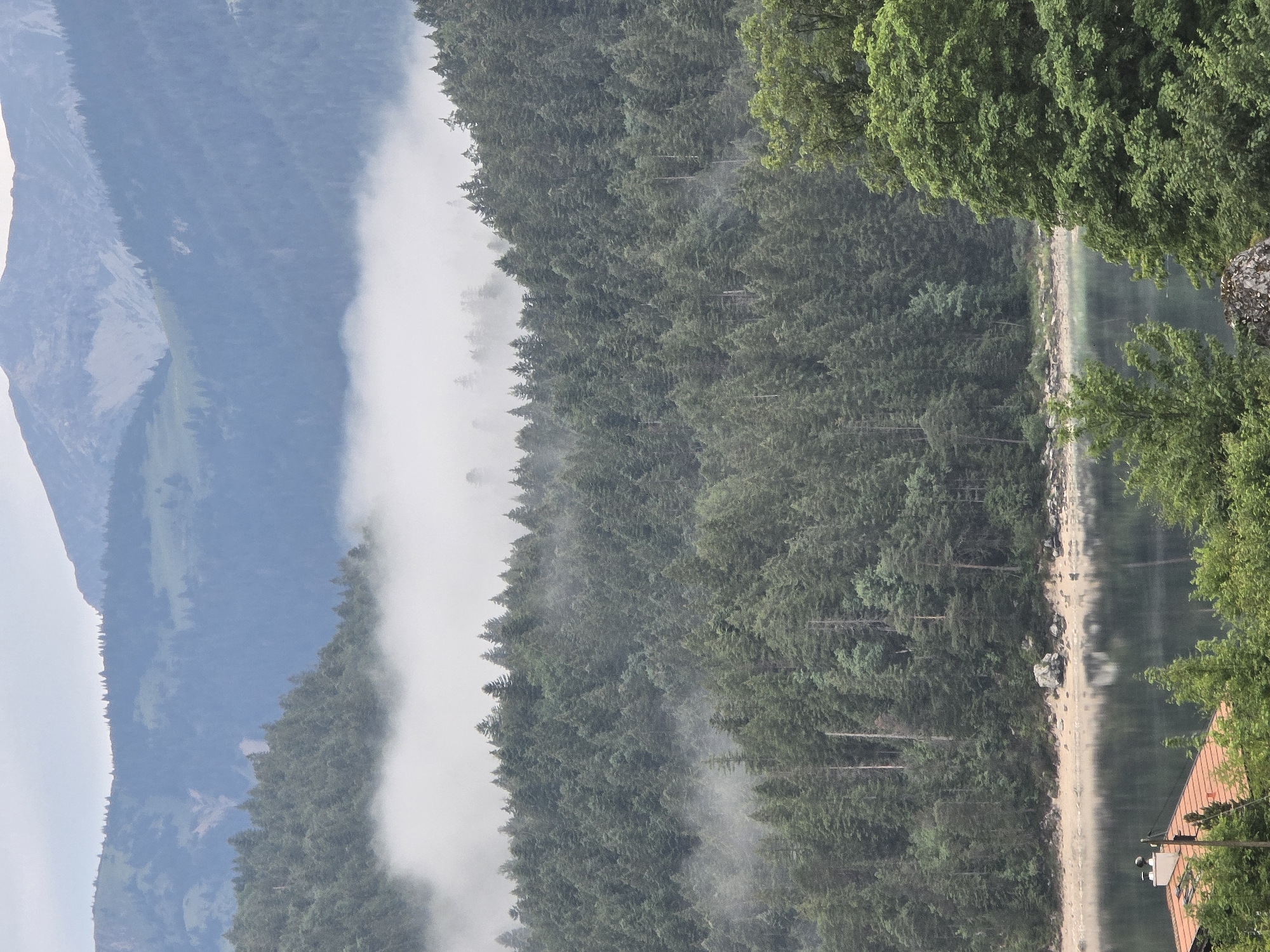 This is an image of a mountainous landscape with a lake in the foreground. The mountains are covered in evergreen trees and there is fog or mist hanging in the valleys. The lake is calm and reflects the trees and mountains. In the lower left corner of the image, there is a red-tiled roof with a white chimney, suggesting a building is present. The scene is likely in or near Grainau, Germany, a town known for its alpine scenery and proximity to lakes and mountains.