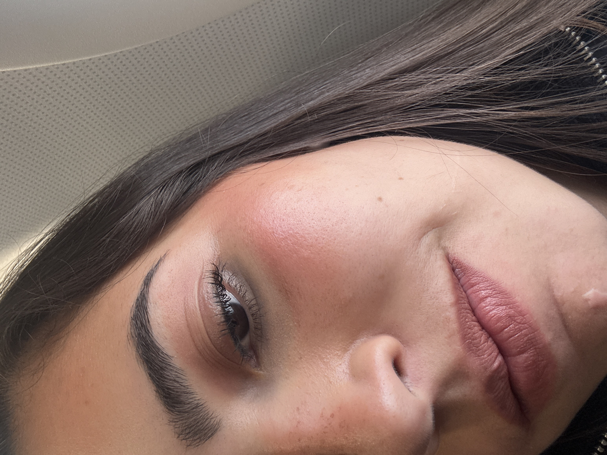 The image is a close-up, angled shot of a woman's face. She has brown hair, fair skin with freckles, and is wearing makeup, including blush, eyebrow filler, and lipstick. Her eye is open and focused. The background appears to be the interior of a car, as indicated by the textured ceiling. The location within San Antonio, United States of America, cannot be determined from the image alone.